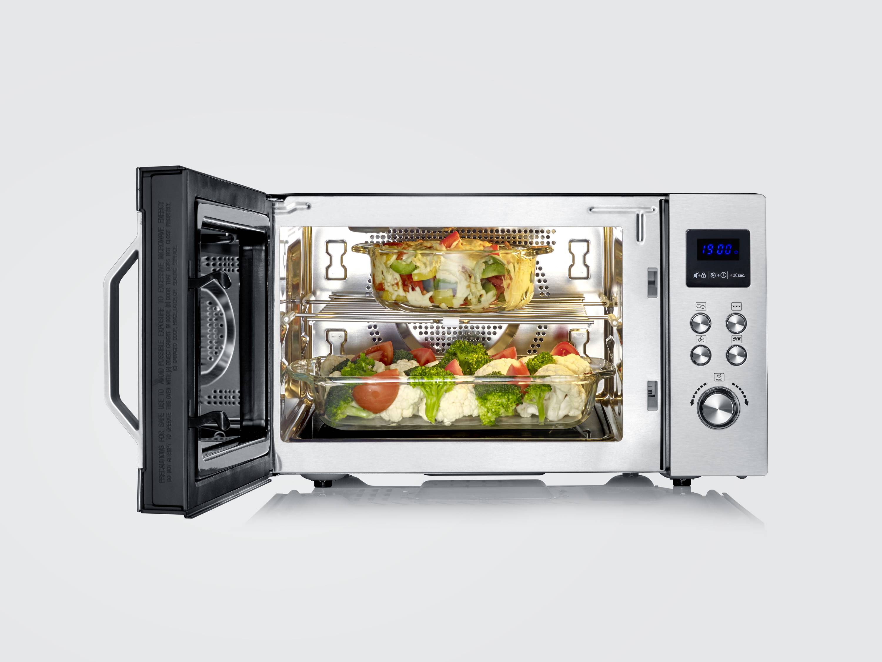 An open hot air oven shows vegetables on two baking trays inside. The control panel and display are located on the right-hand side.