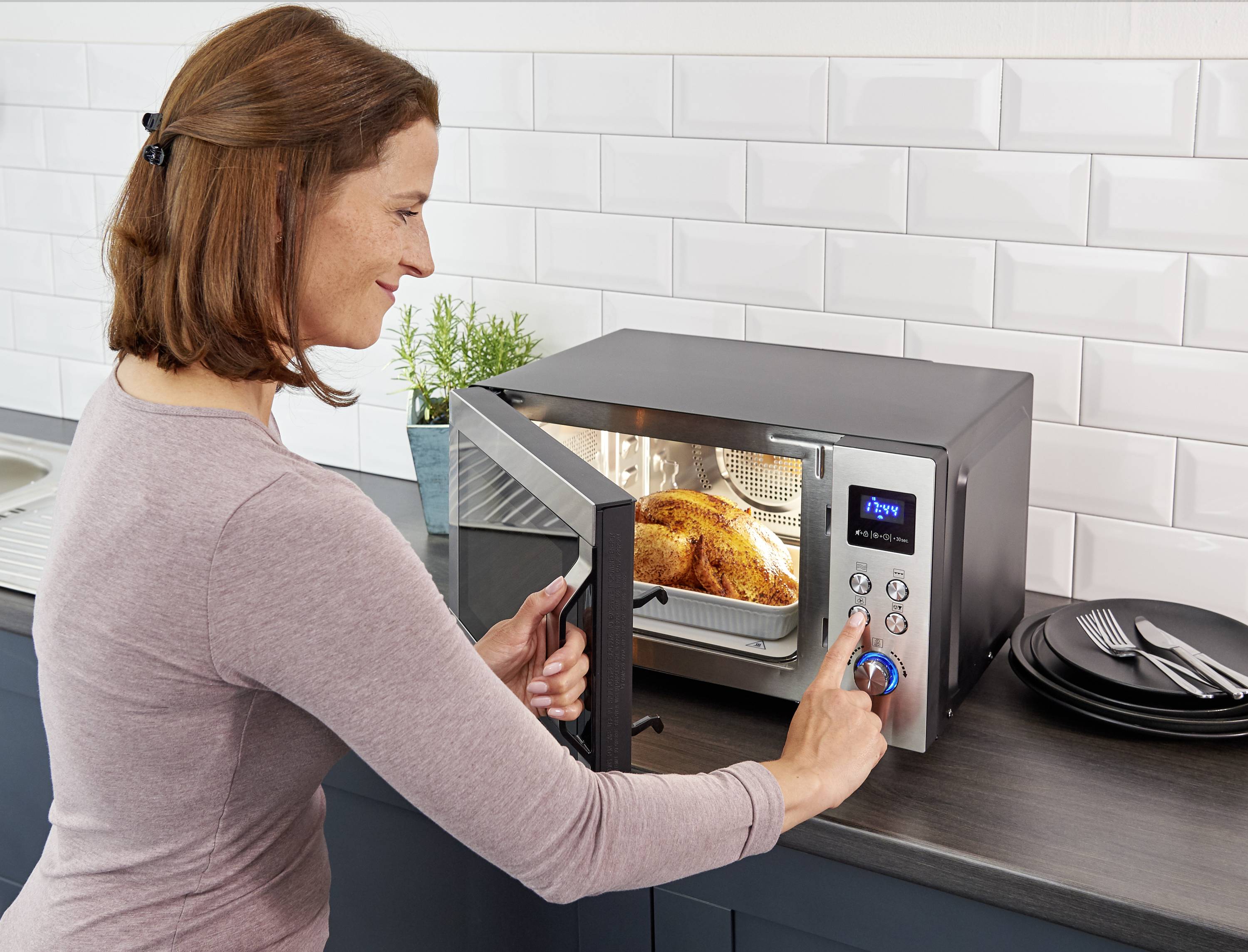 A woman opens a microwave containing a roasted chicken. She presses the buttons to operate the appliance.