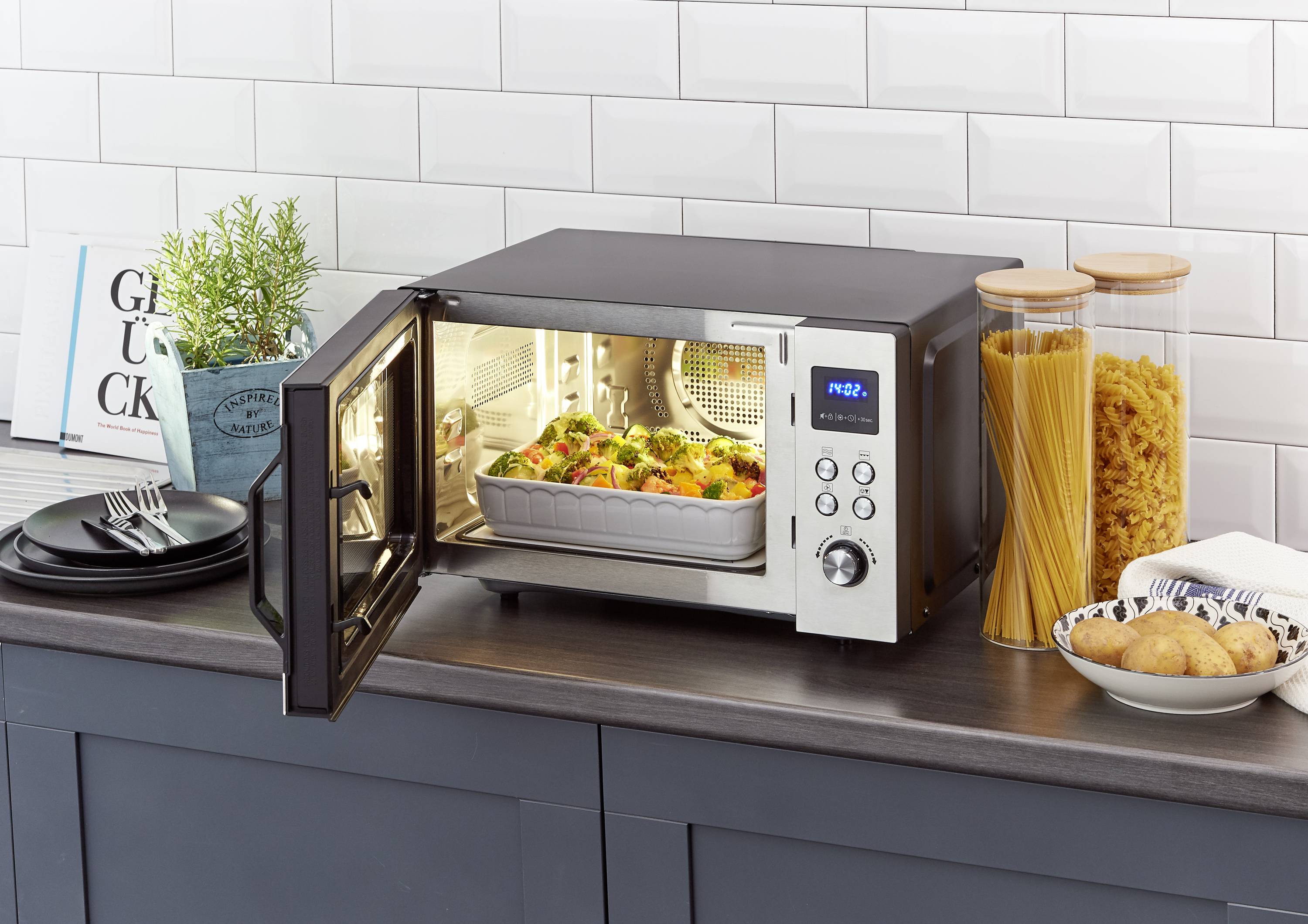 An open microwave on a kitchen worktop, filled with a casserole. Next to it are glass containers with pasta and several plates.