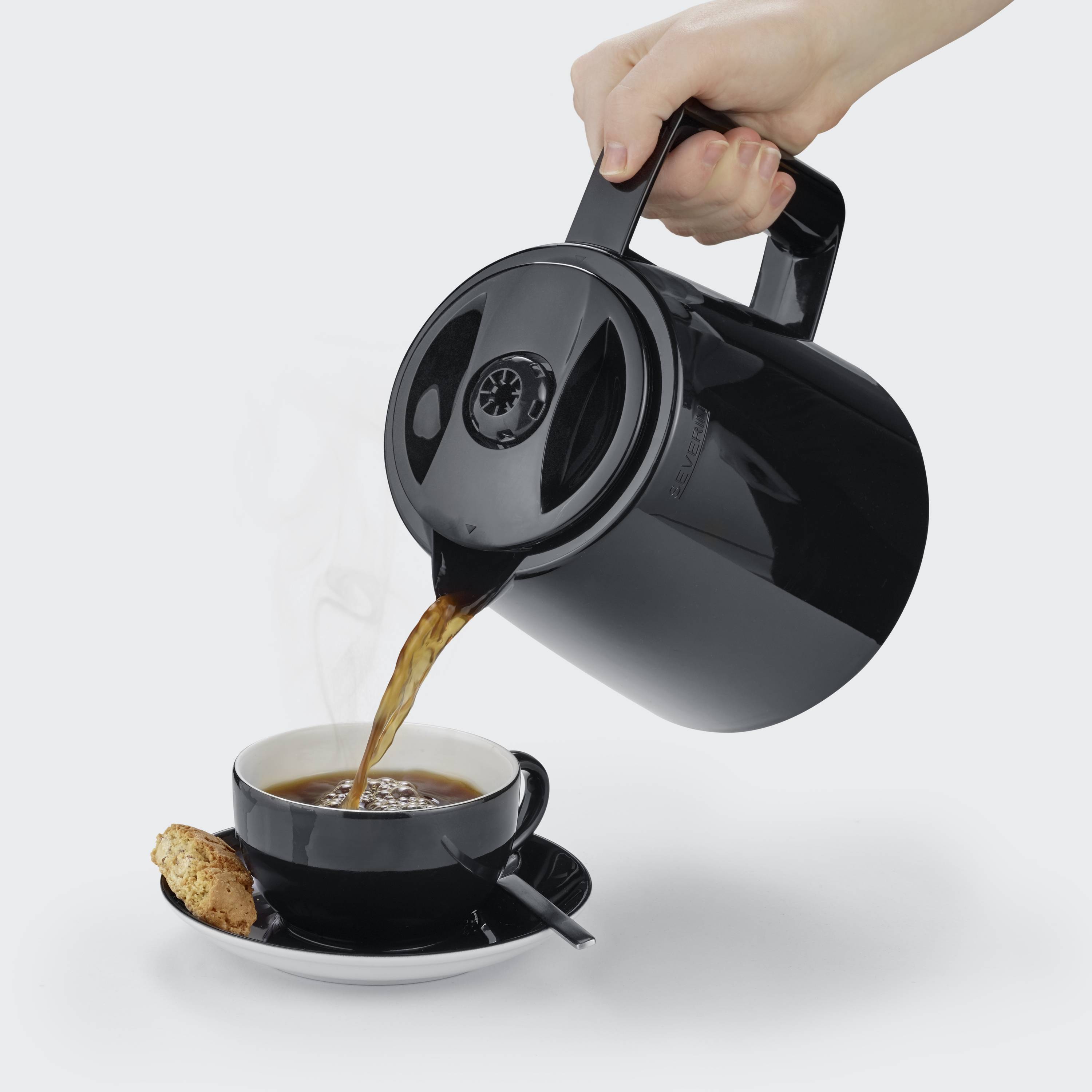 A hand is pouring coffee from a black jug into a cup, which is sitting on a saucer with a biscuit and a small spoon.