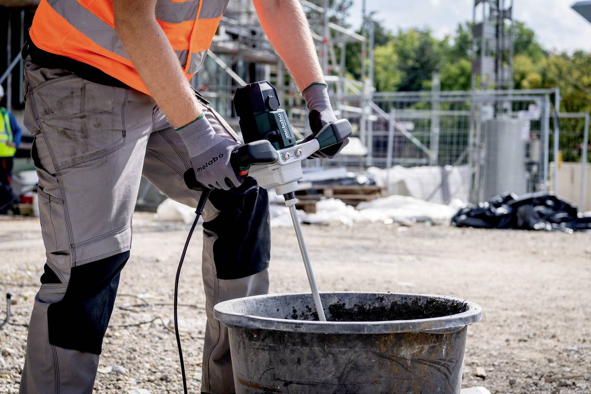 A construction worker is mixing mortar in a bucket on a building site. He is wearing an orange high-visibility vest and grey work gloves.