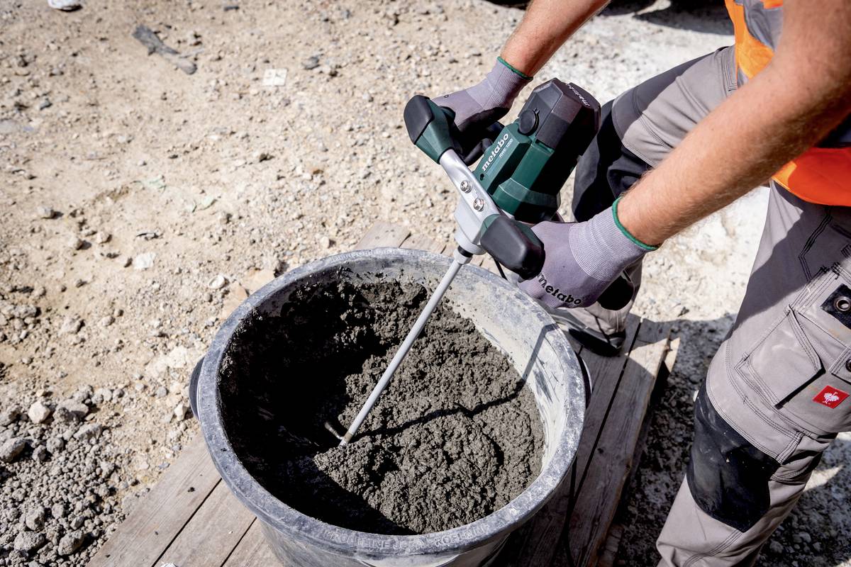 A person is mixing cement in a bucket with an electric mixer. They are wearing work gloves and an orange protective suit.