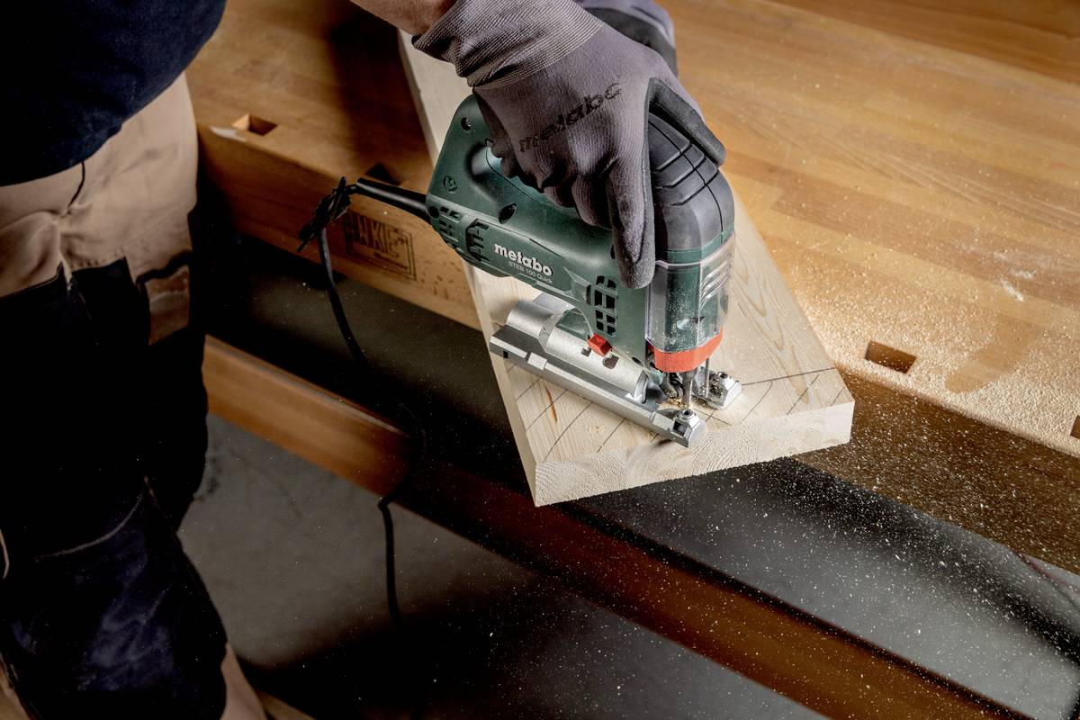 A person is cutting a piece of wood with a jigsaw in a workshop. Sawdust is visible, indicating active work in progress.