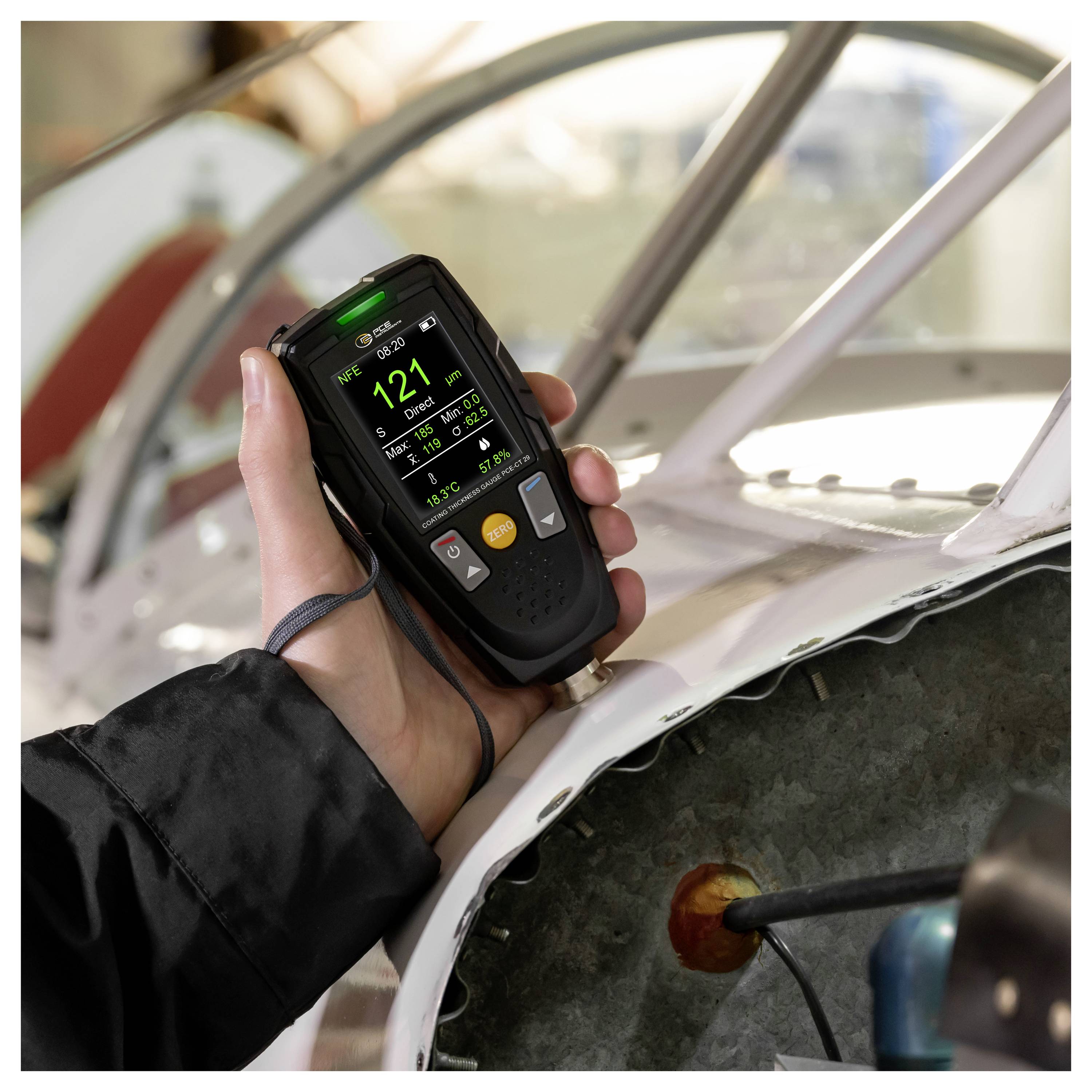 Hand holding a digital inspection meter against an exposed metal surface, measuring a white vehicle or boat hull during repair or testing.