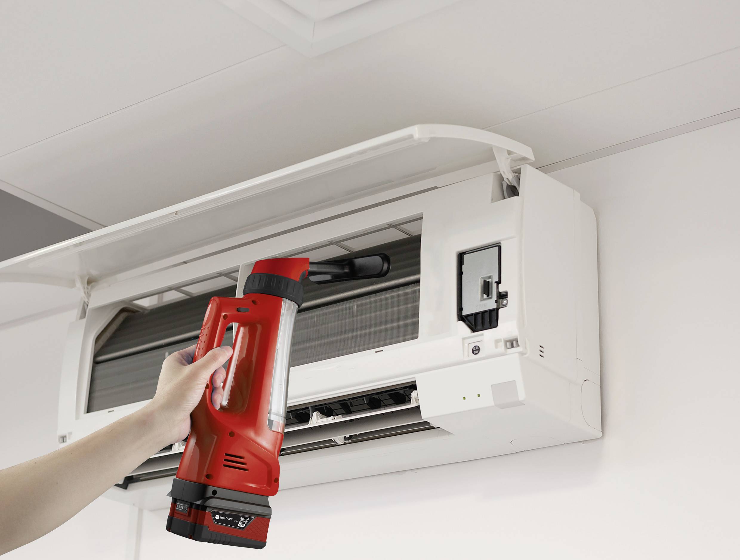 A hand is holding a red and black device in front of an open air conditioning unit for cleaning or inspection. A ceiling is visible.