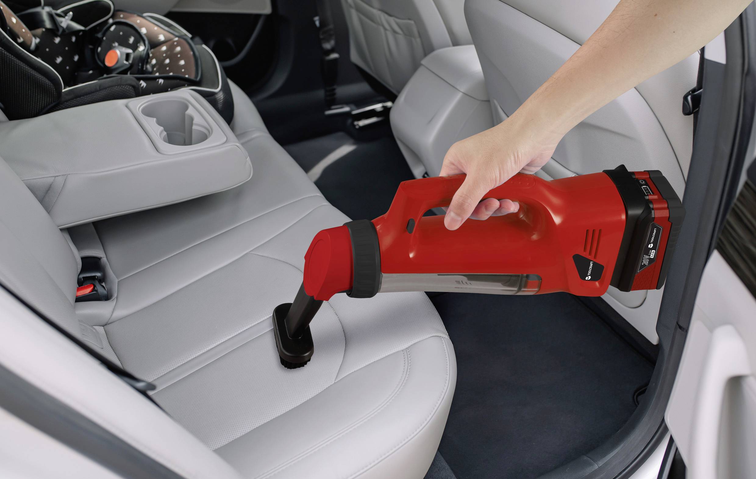 A hand is cleaning the rear seats of a car with a red cordless vacuum cleaner. A child seat is positioned on the back seat.