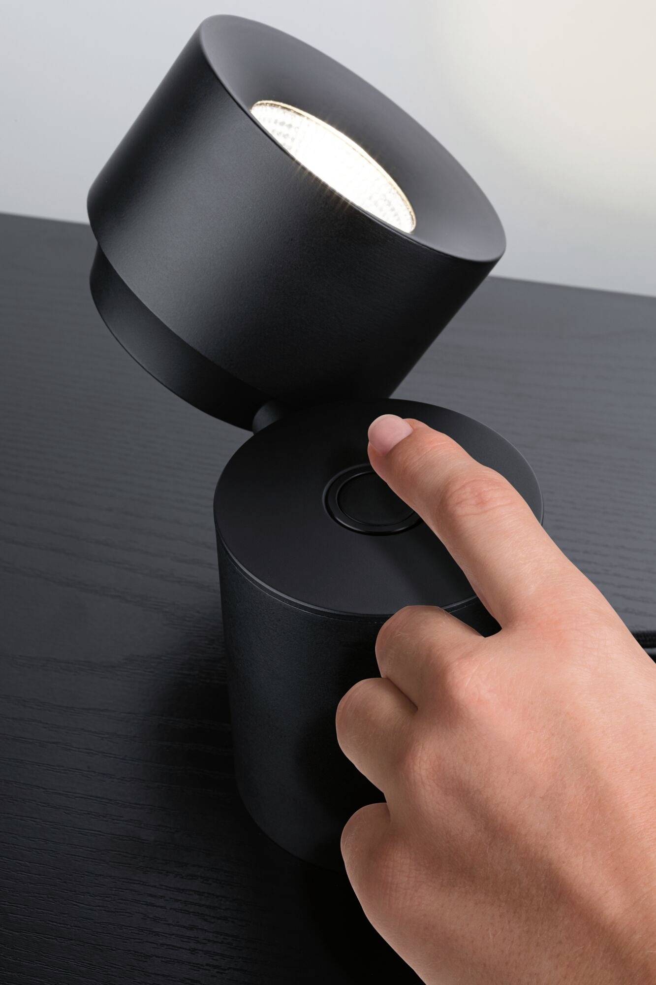 A hand presses a switch on a modern, black lamp situated on a table. The lamp is adjustable and provides illumination.