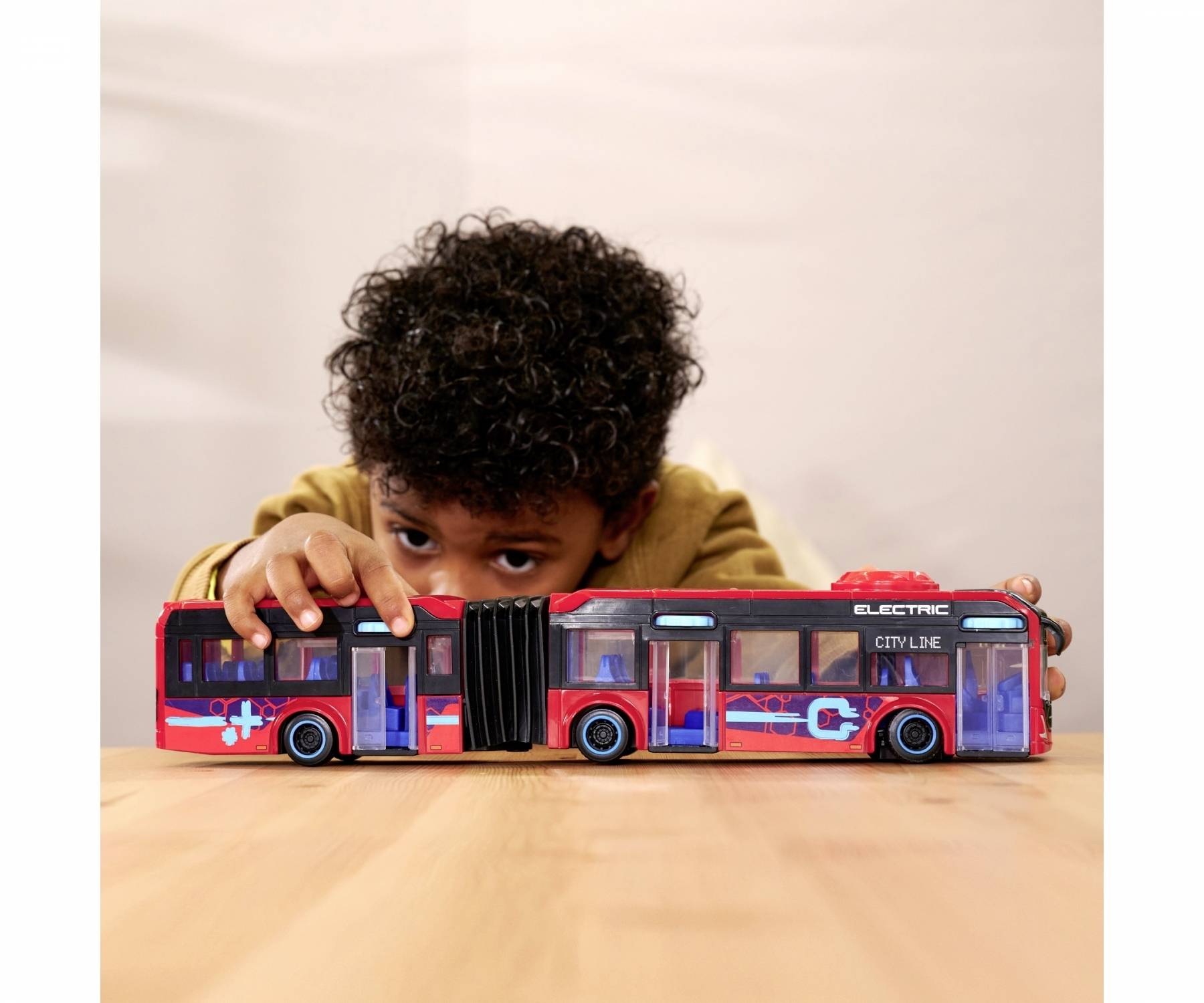 A child is playing with a large model bus on a table. The bus is red with blue and white details and bears the inscription 'ELECTRIC'.