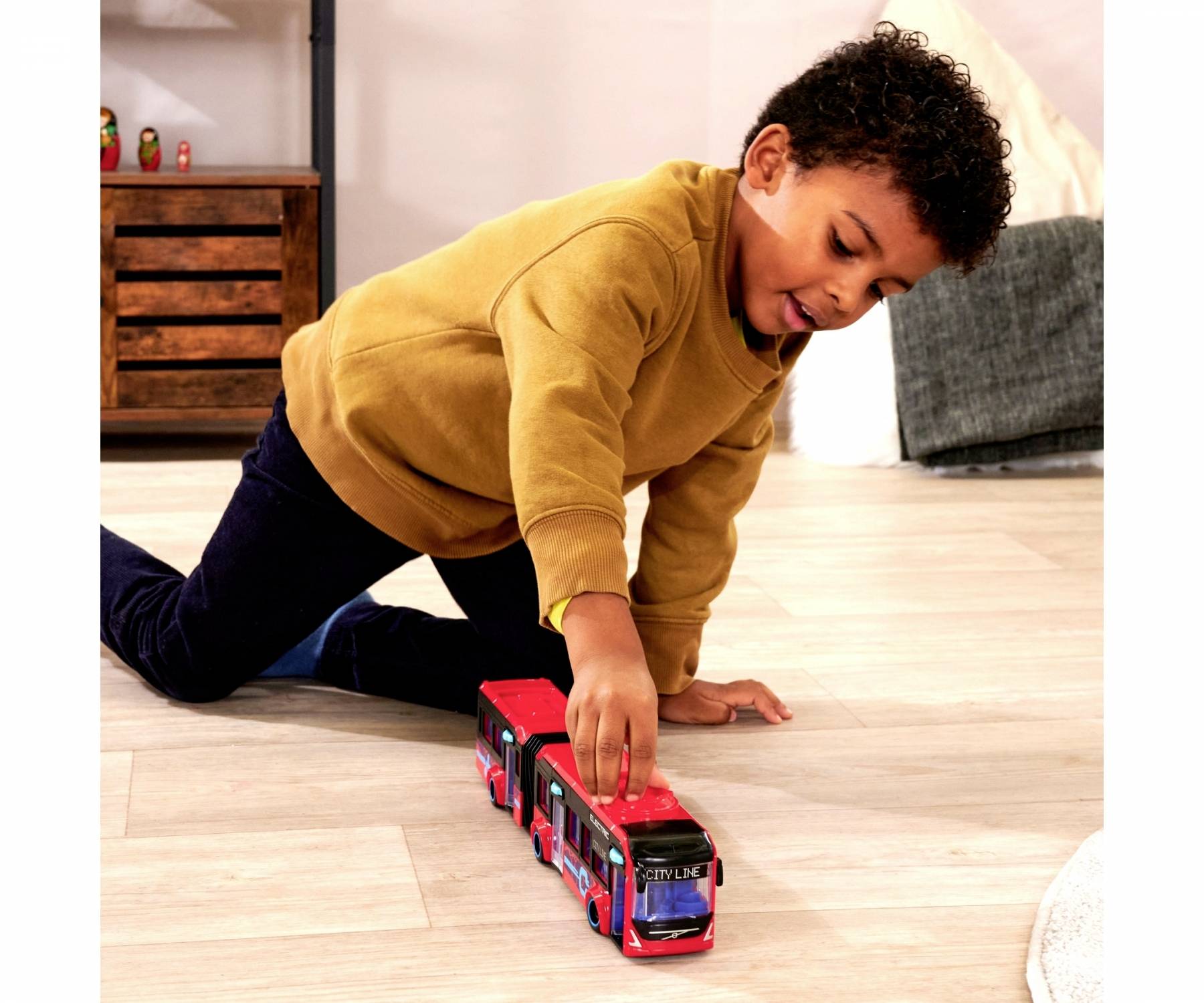 A child is playing with a red toy bus on a wooden floor. They are wearing a yellow jumper and blue jeans.