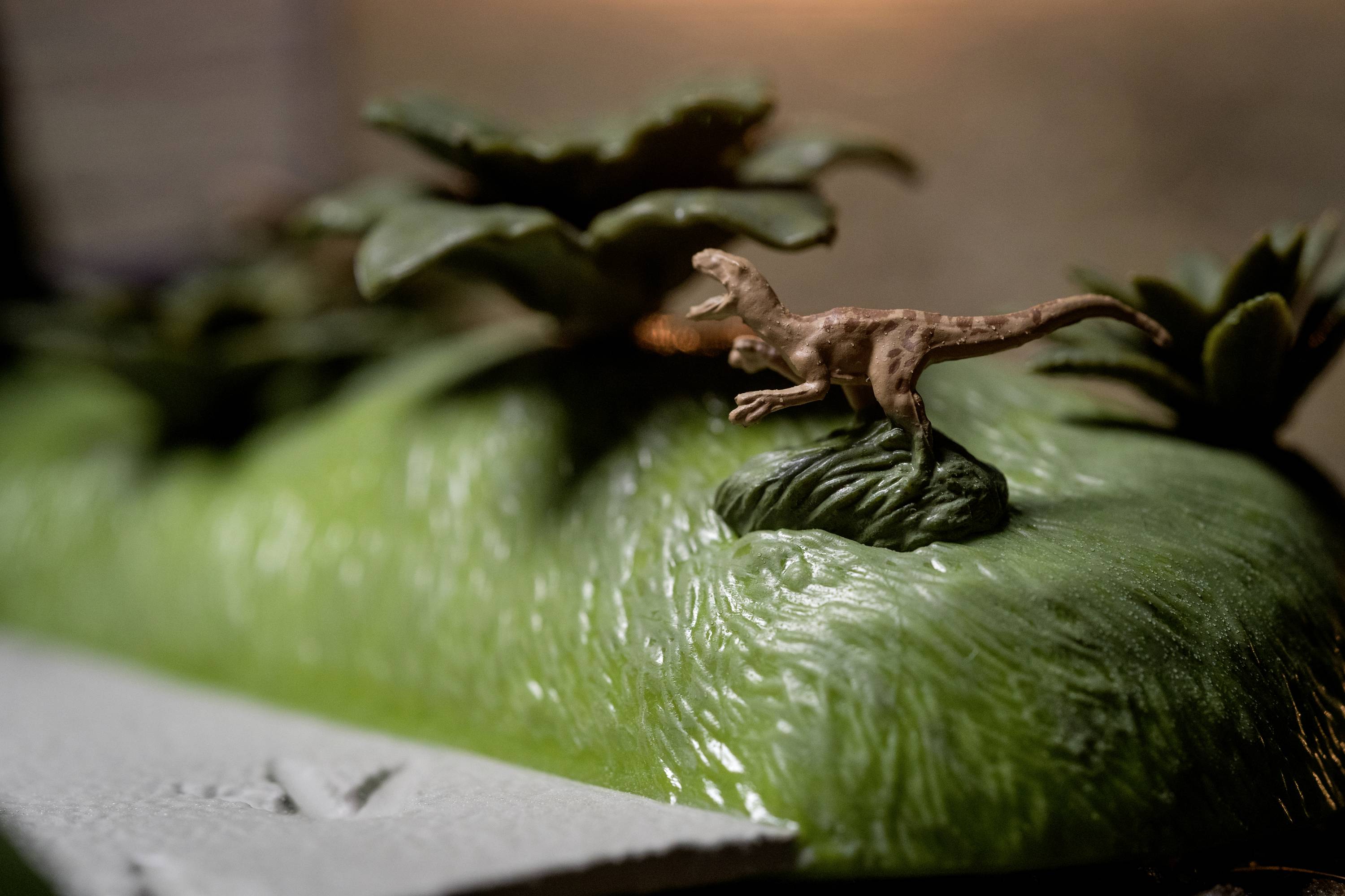 A small plastic model of a dinosaur stands on artificial grass, with plastic plants visible in the background.