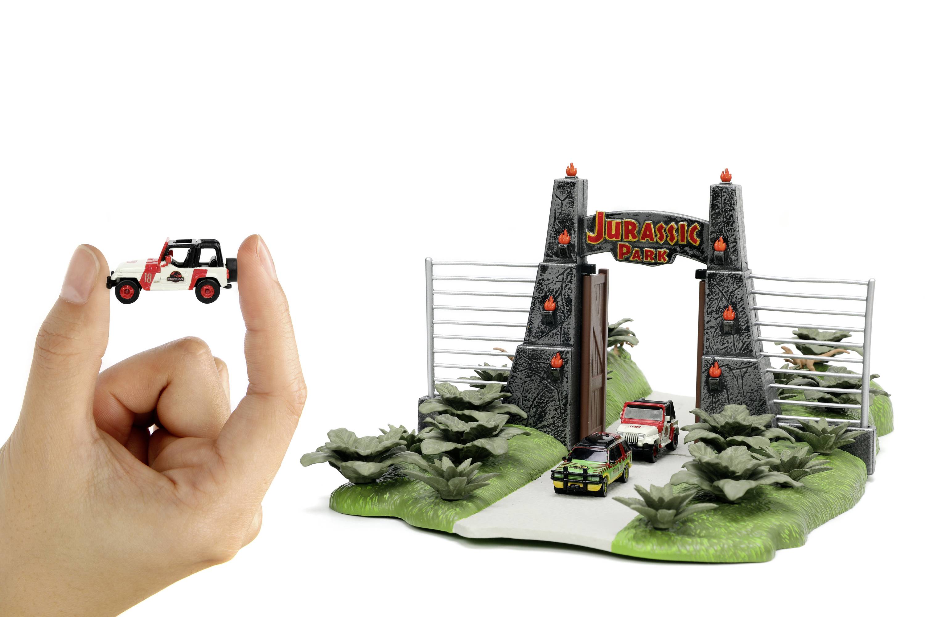 A hand holds a small toy car in front of a model of the 'Jurassic Park' entrance gate, with two miniature cars positioned in front of it.