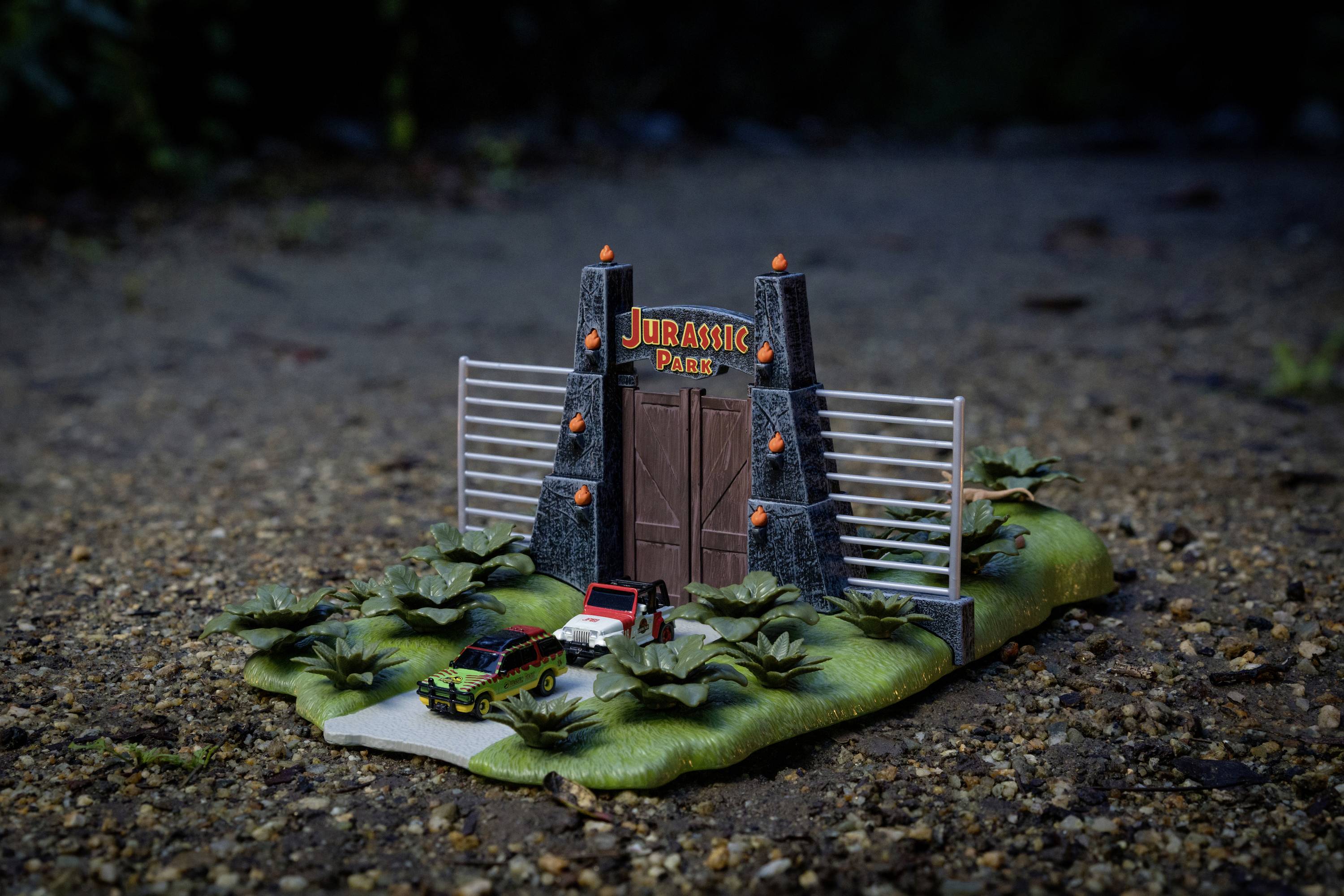 Miniature replica of the 'Jurassic Park' entrance with two toy cars in front, surrounded by plants, on a sandy surface.