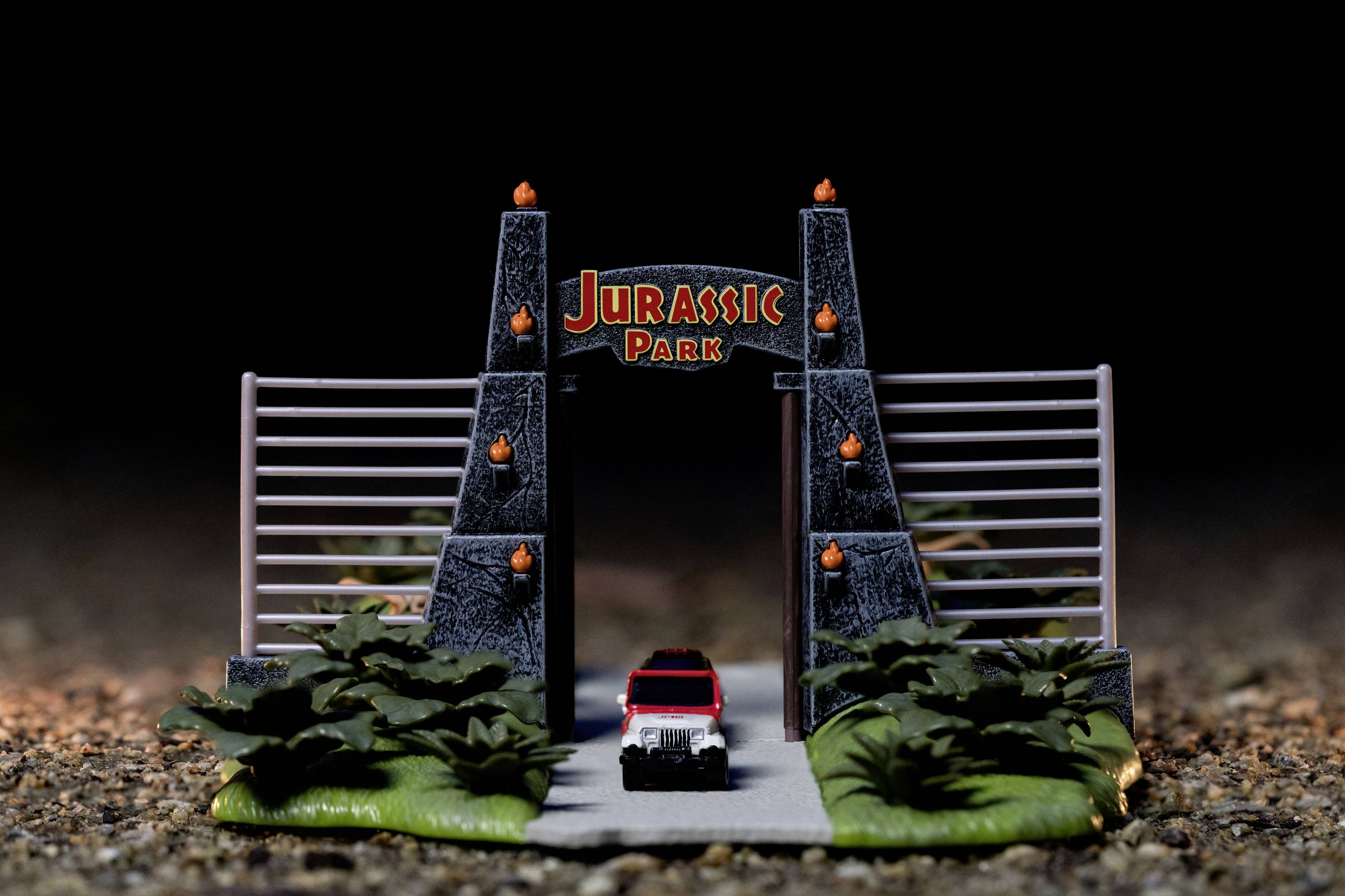 A small car drives through a miniature gate with the 'Jurassic Park' inscription, surrounded by plants, on a rocky surface.