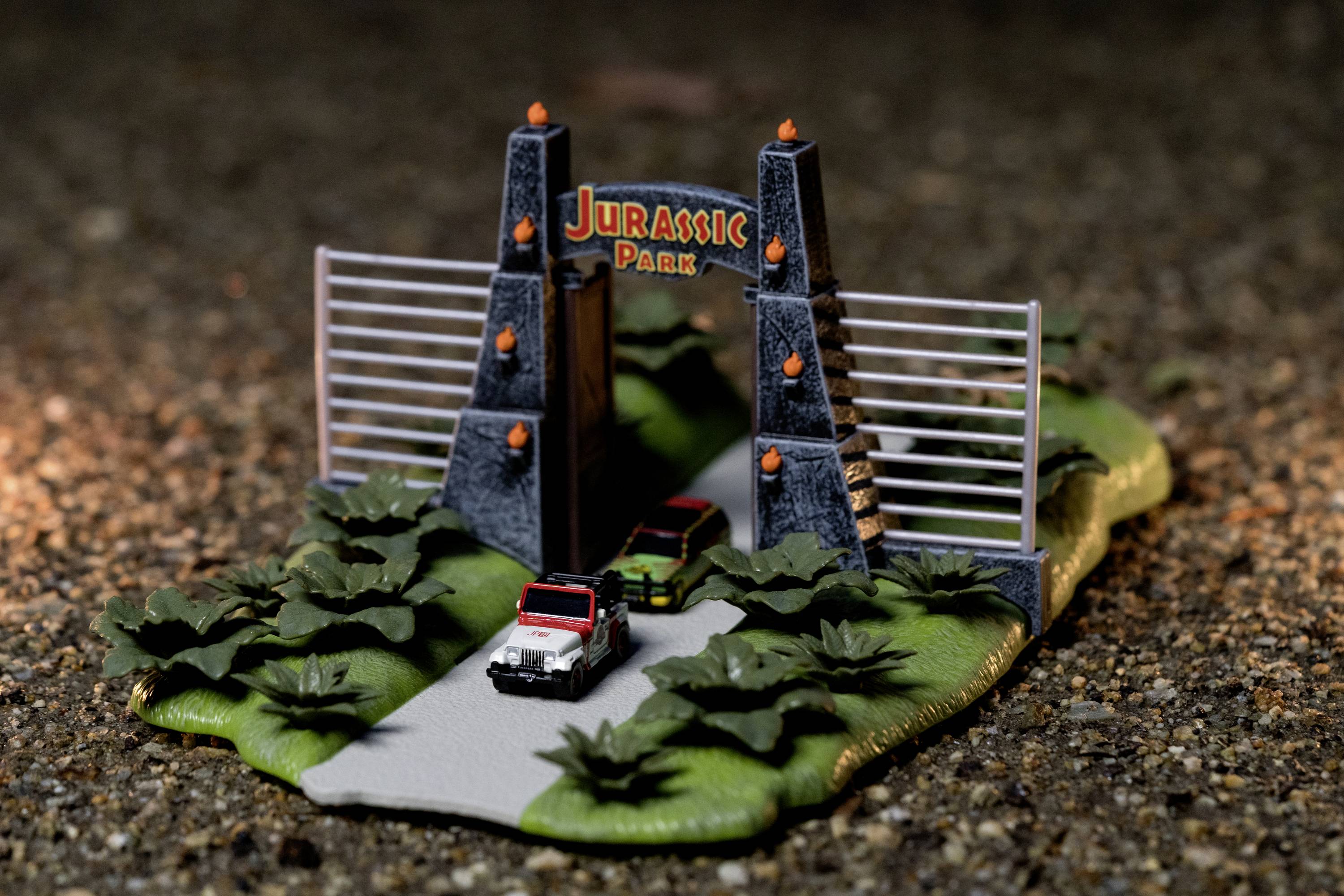 Miniature model of Jurassic Park featuring two vehicles, the gate, and surrounding landscape. The vehicles are parked facing the gate.
