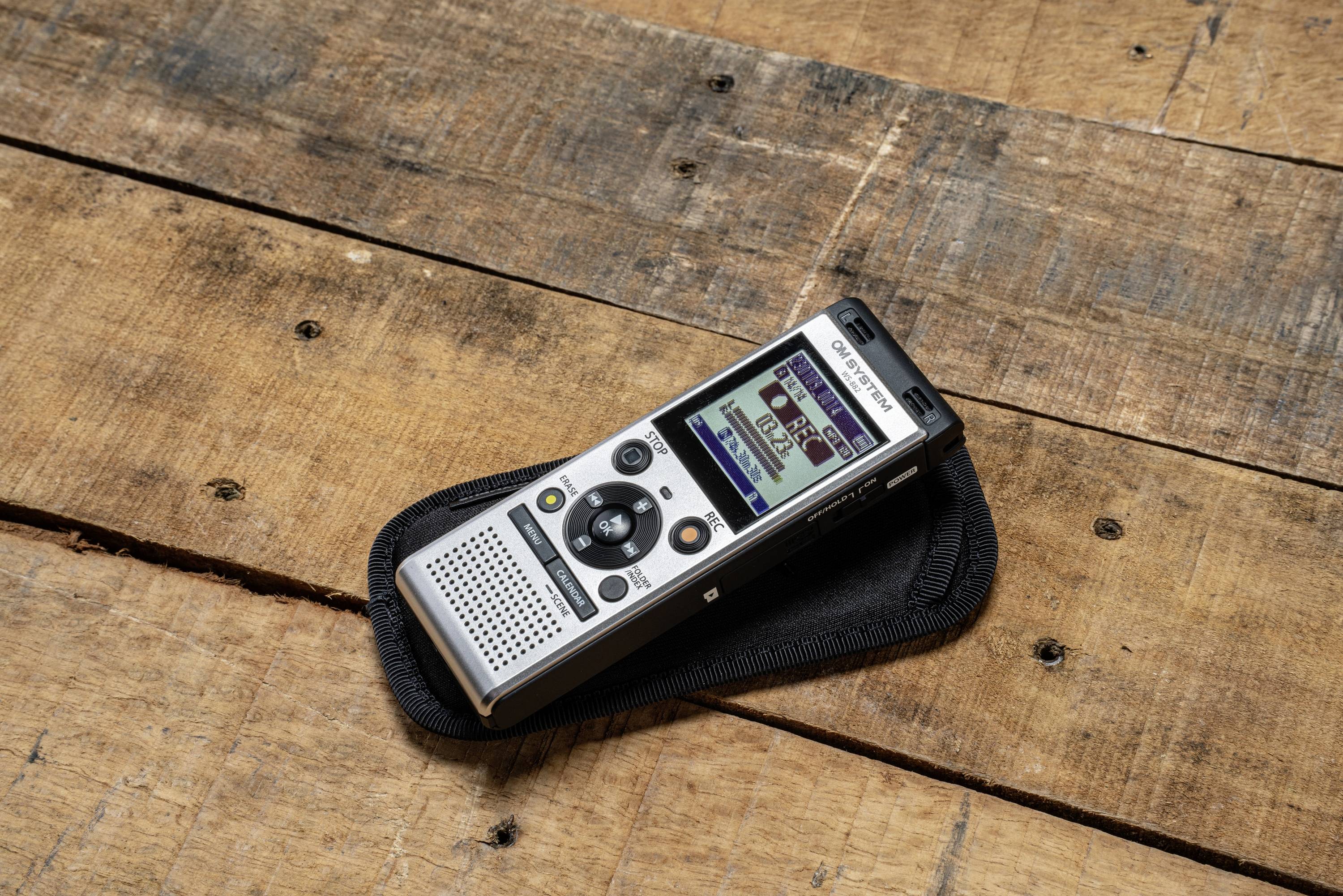 Portable dictation device with display, resting on a wooden table. Device shows recording function, used for audio recordings.