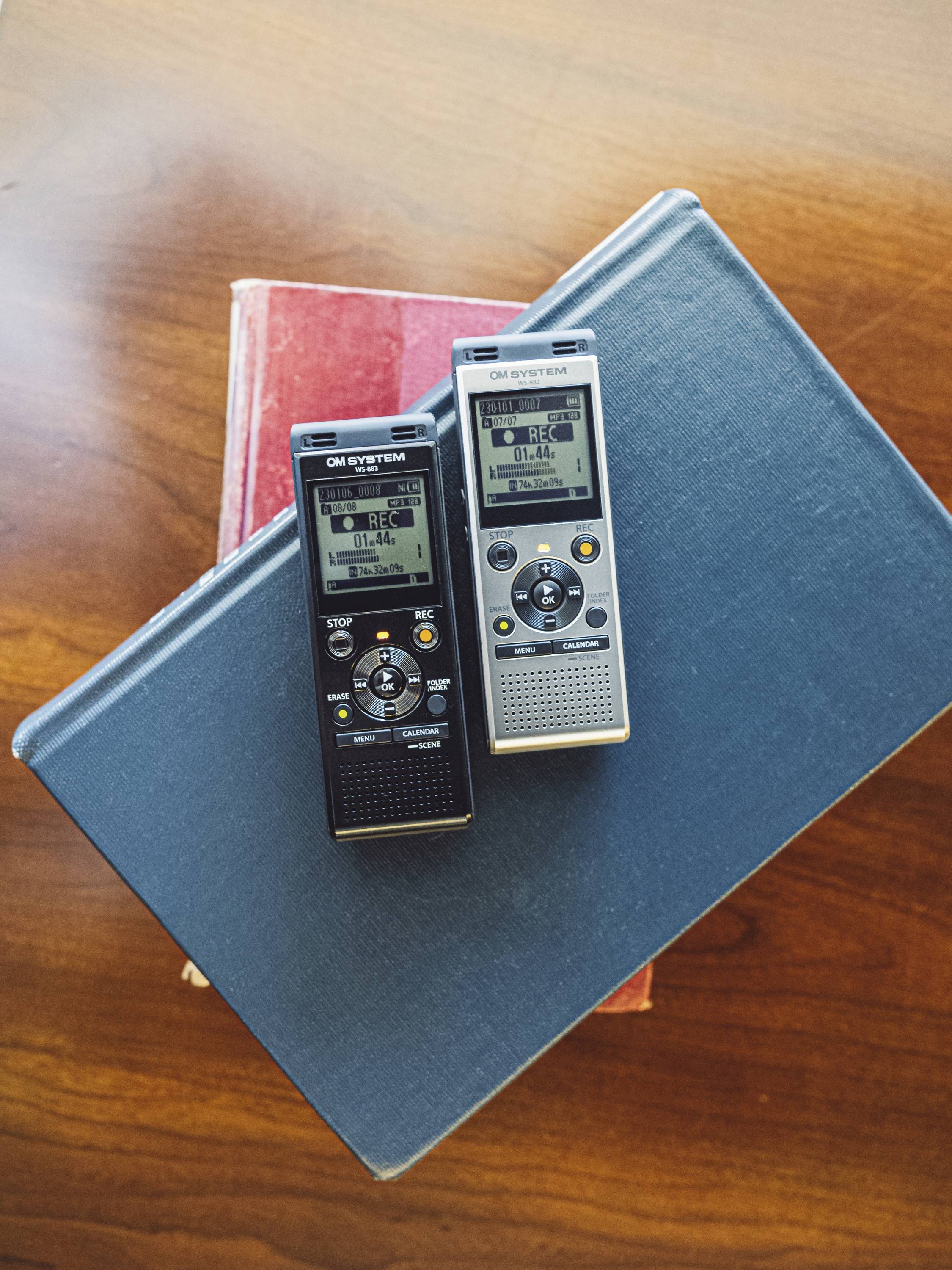 Two digital dictaphones lie on a stack of books with a wooden table in the background.