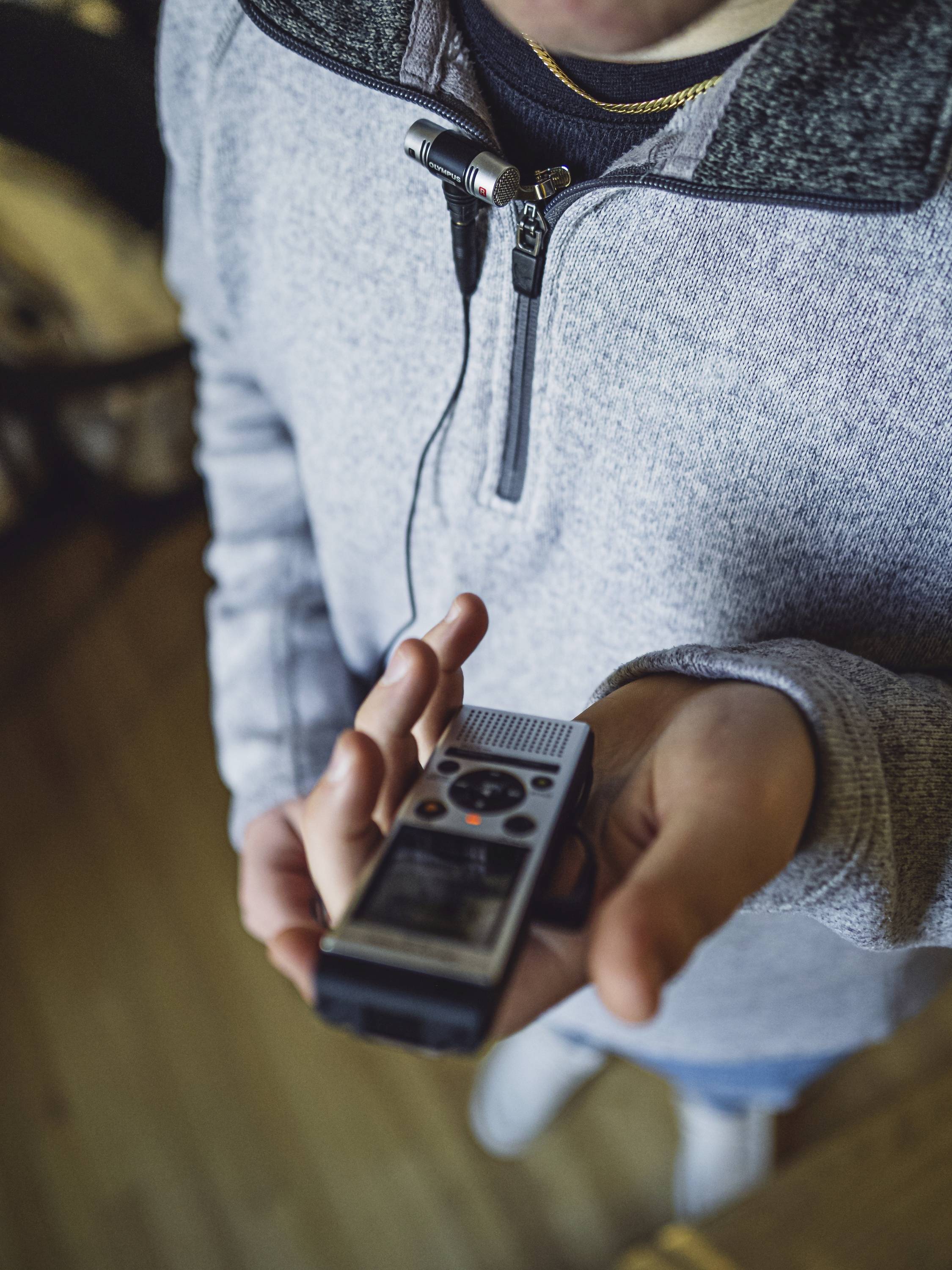 A person is holding a recording device in their hand, with a microphone attached to a grey jumper. Focus on the dictaphone.