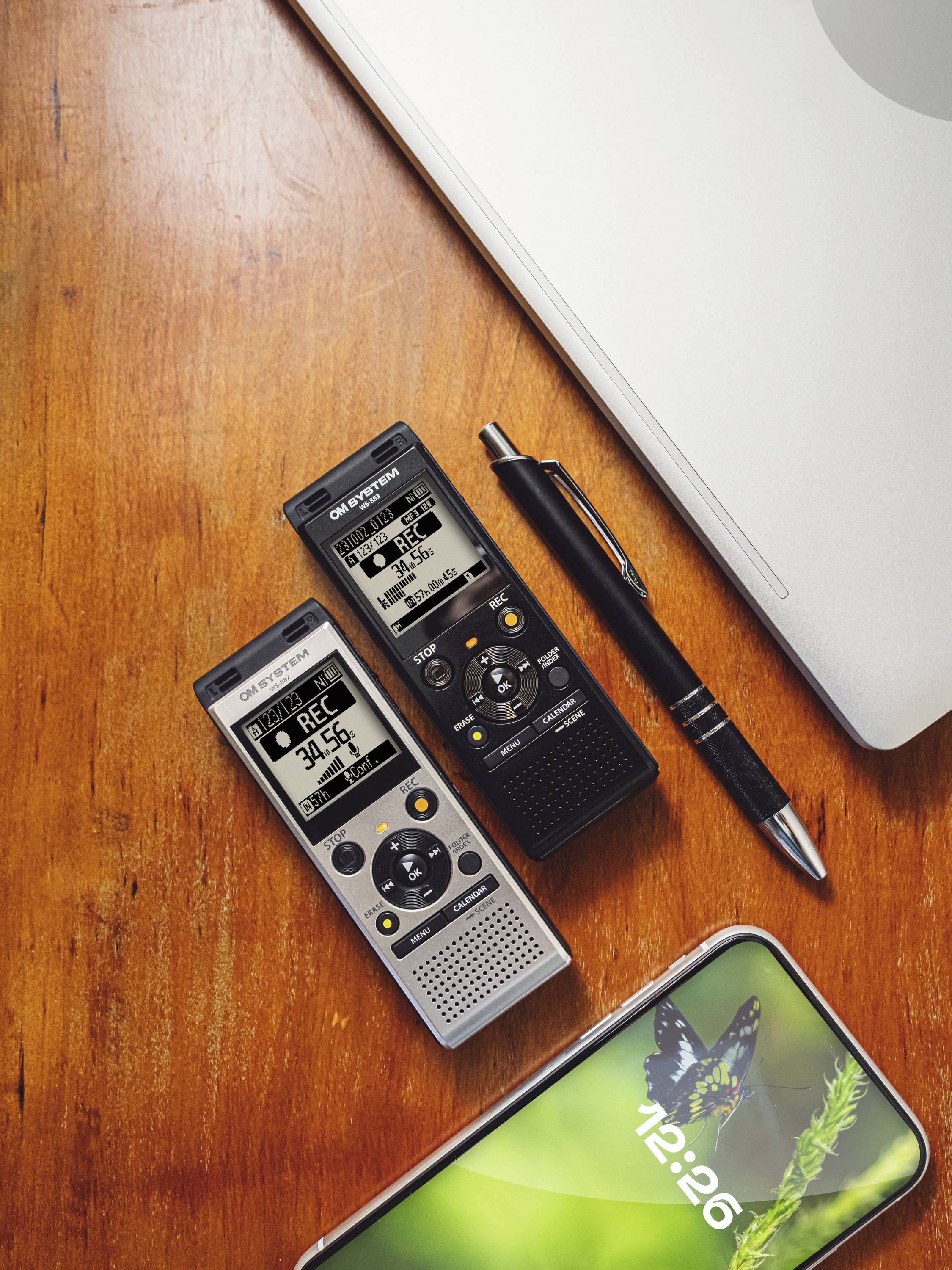 Two digital dictaphones, a pen and a smartphone on a wooden table next to a laptop. The smartphone displays an image of a butterfly.