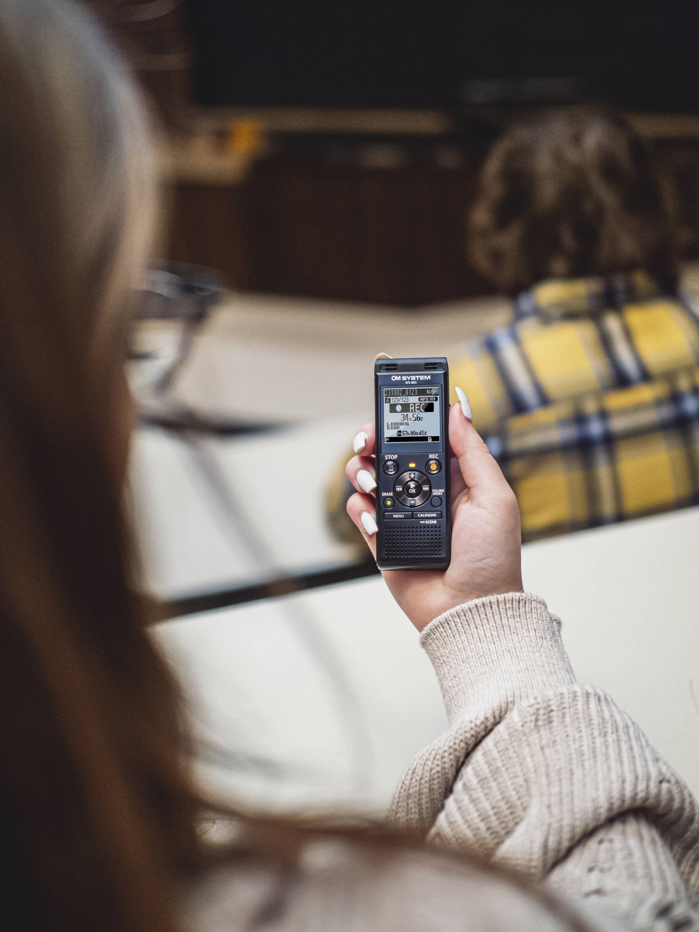 A person is holding a digital audio recorder in their hand. In the background, another person sits blurred at a table.