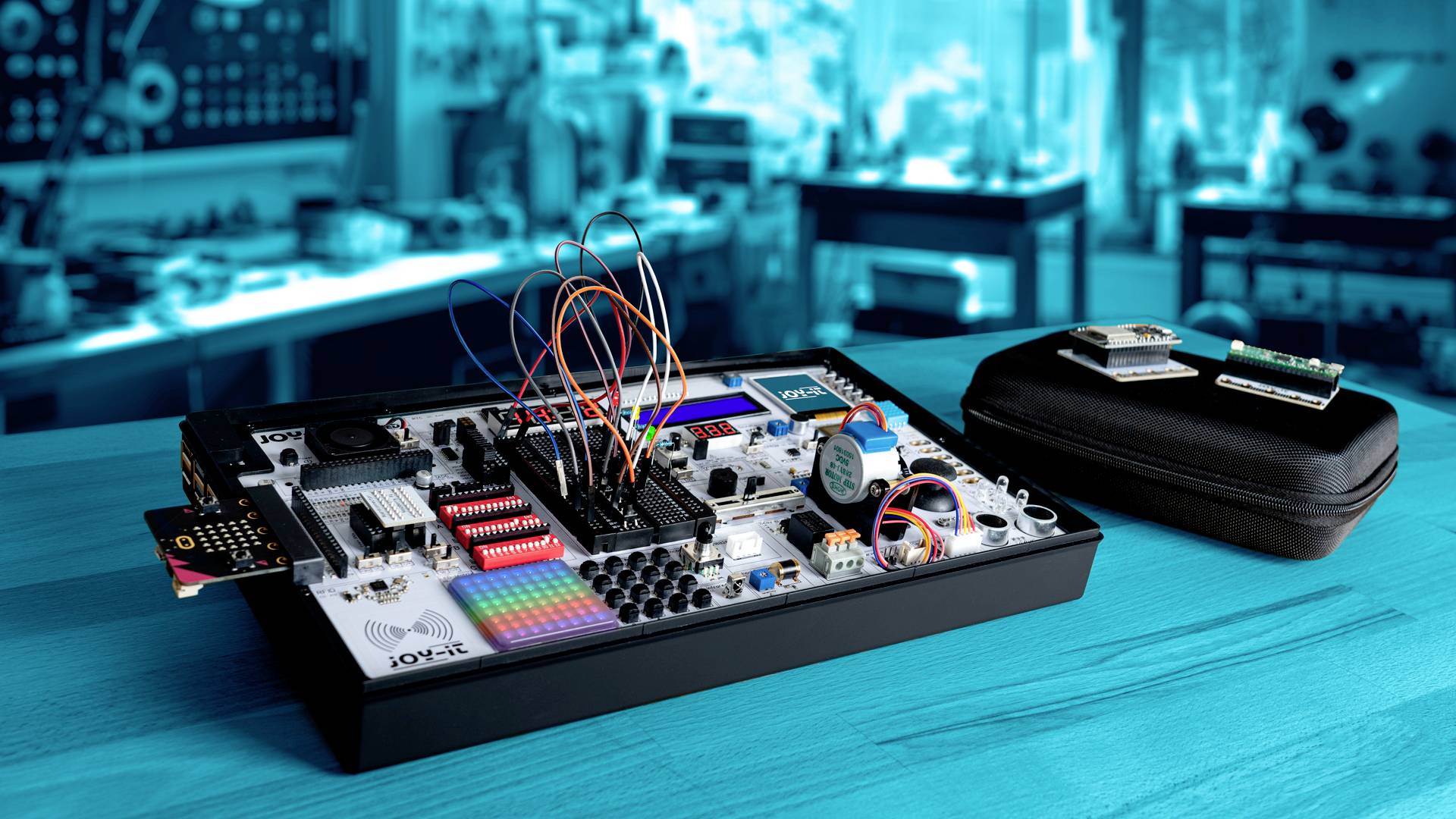 Electronic kit with cables and components on a wooden table, blurred workshop background; shows learning equipment for electrical engineering.