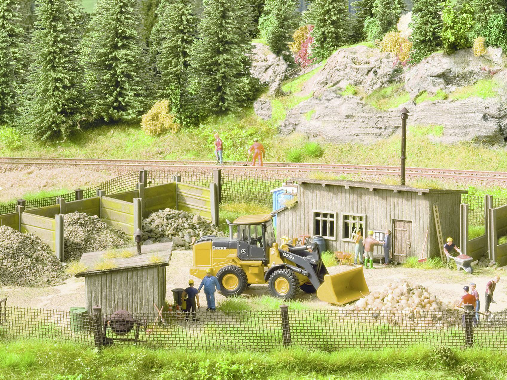 A model-making diorama depicts a construction site with an excavator, workers, a wooden hut, and surrounding trees and rocks.