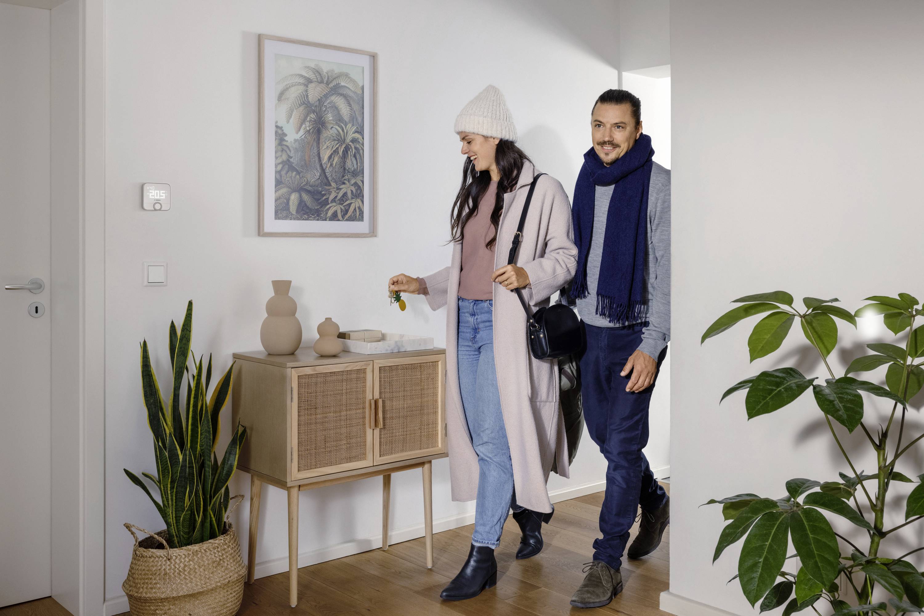 A man and a woman enter a room smiling, both wearing winter clothing. Plants and a wardrobe can be seen in the background.