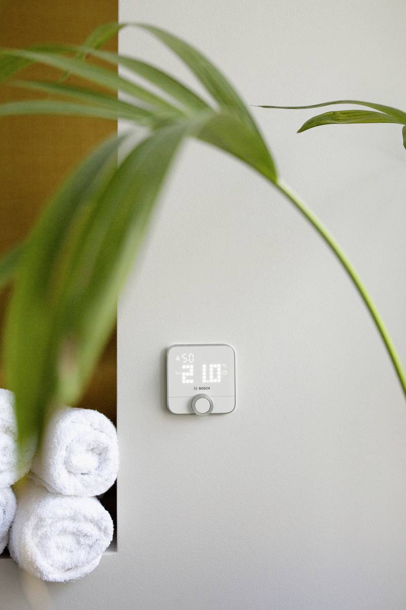 A digital thermostat displays the temperature. In the foreground, a leaf of a houseplant, with rolled-up towels beside it.