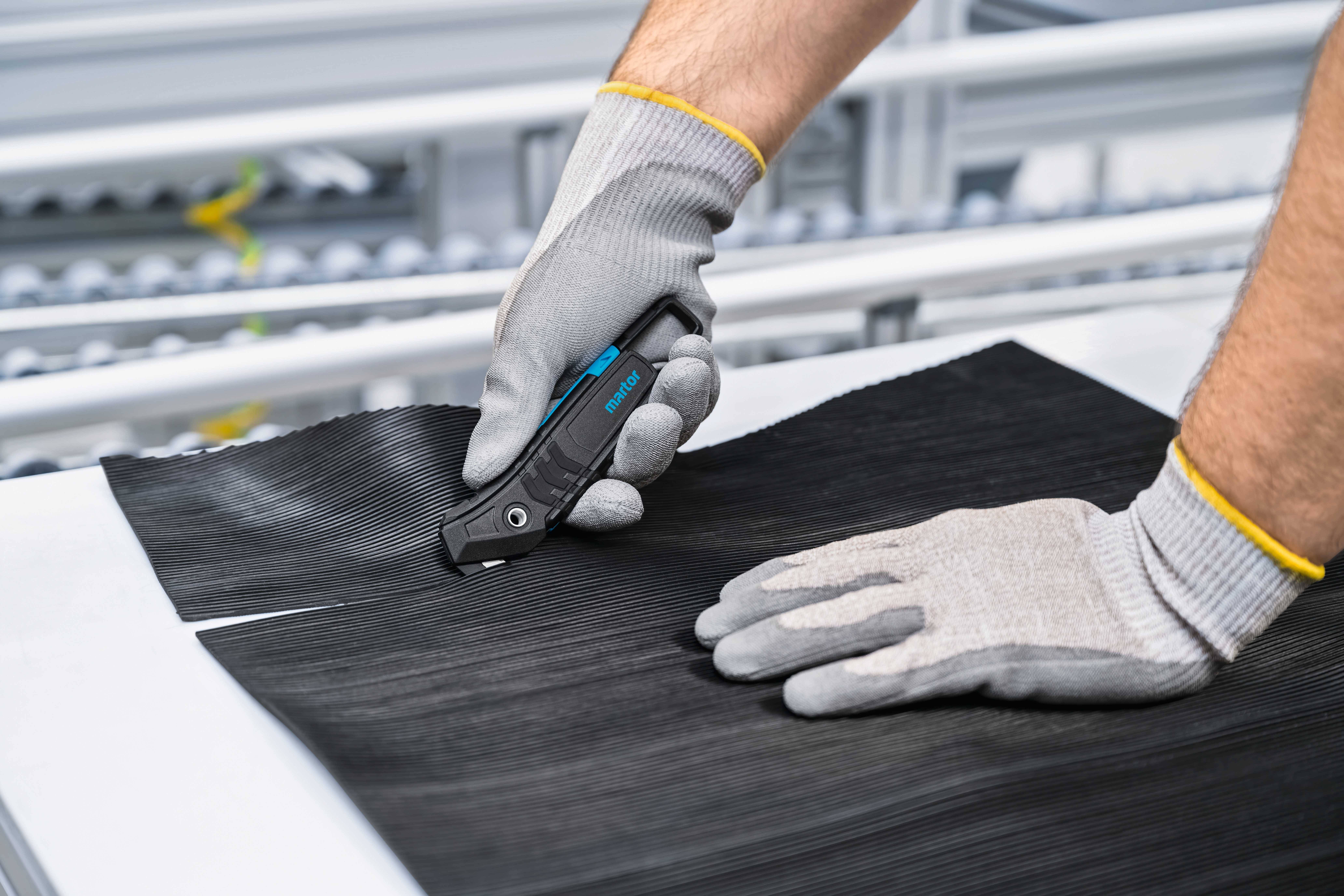 A person is cutting a black material with a safety knife on a table, with both hands protected by grey gloves.