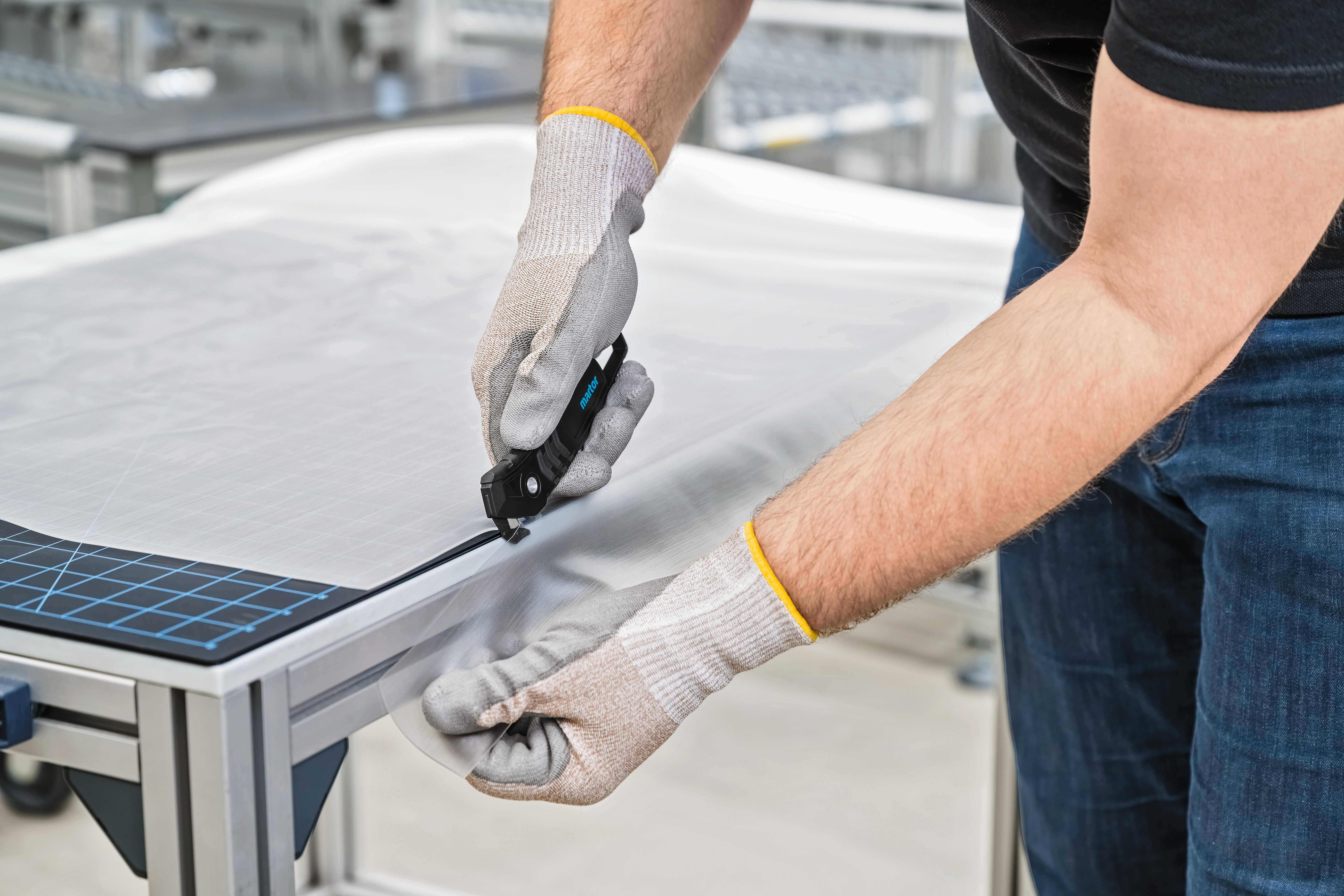 A person carefully cuts fabric on a table using a craft knife, wearing work gloves.