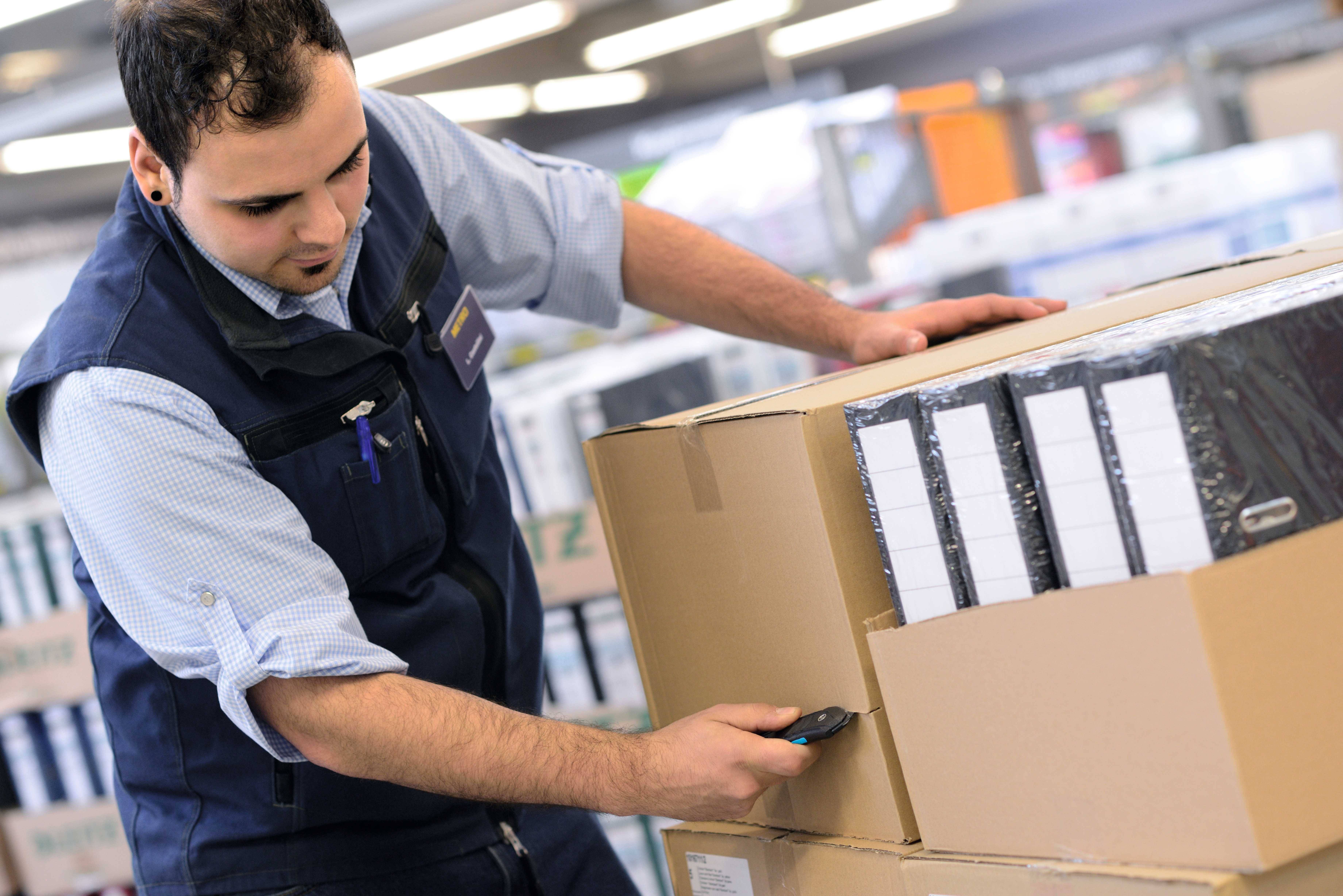 A person is scanning large cardboard boxes with a device in a warehouse. They are wearing a blue hi-vis vest and are standing near shelves in the background.
