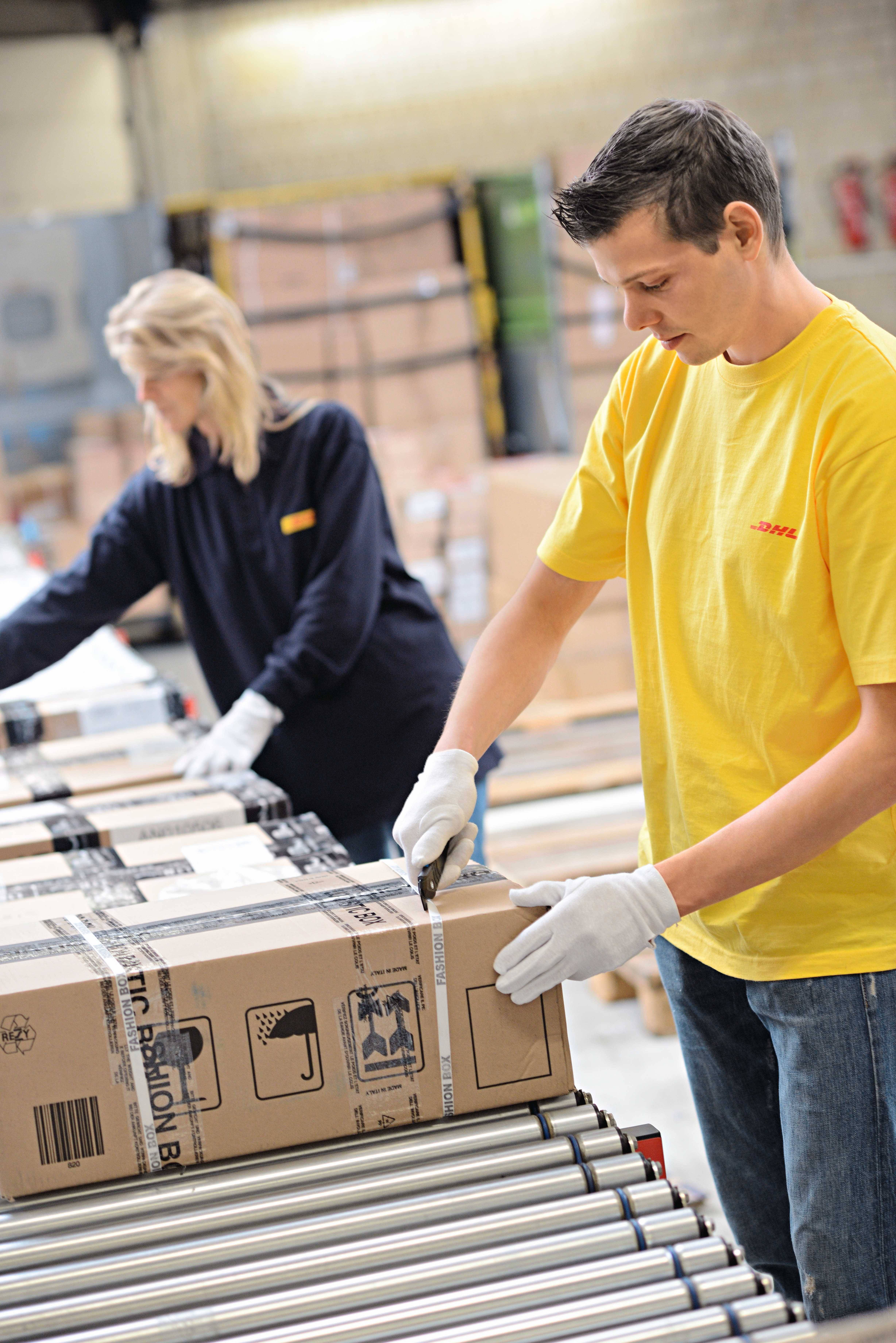 A man and a woman are standing at a conveyor belt packaging boxes in a storage warehouse. Both are wearing work gloves and workwear.