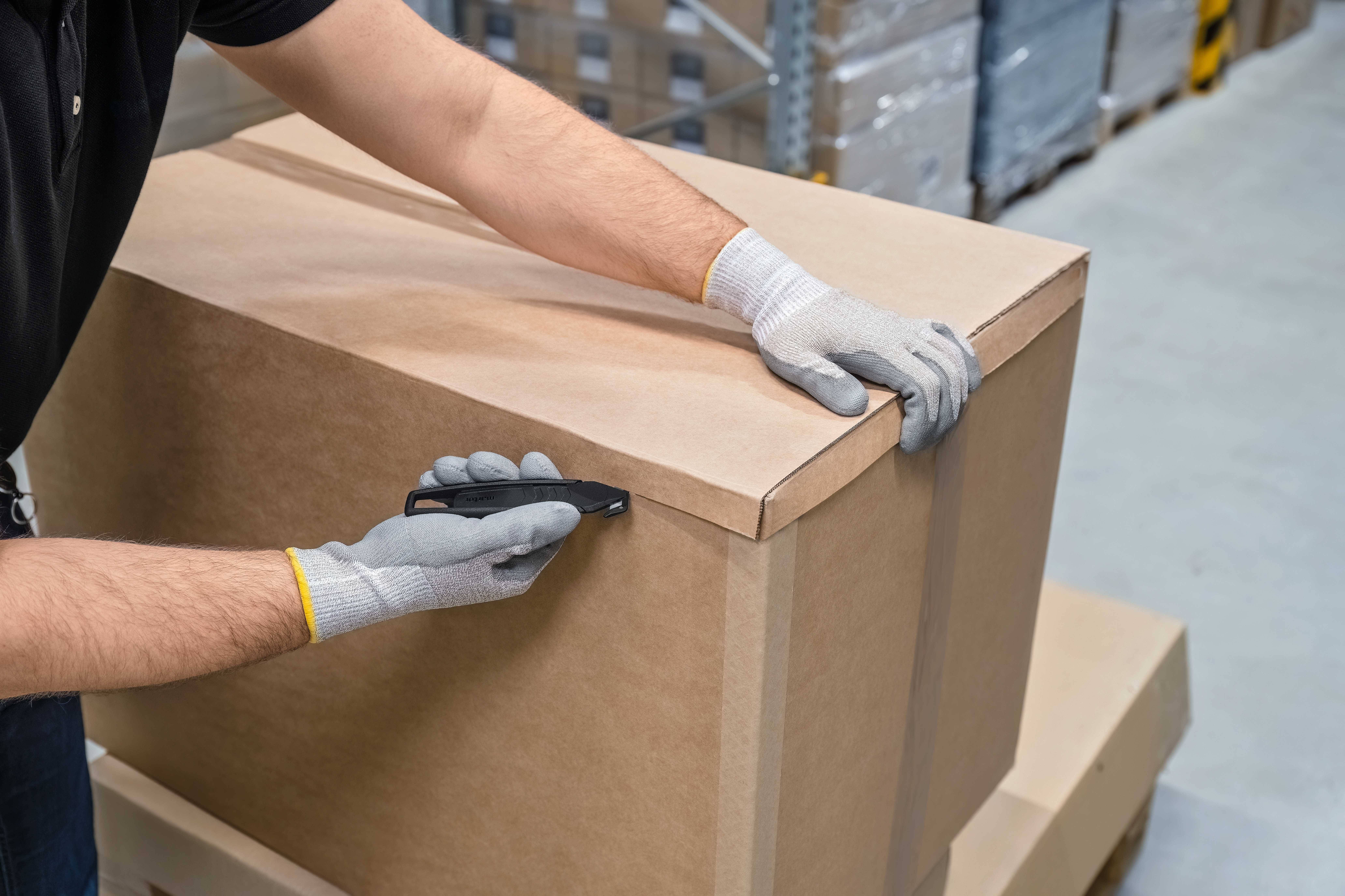 A person opens a large, brown cardboard box with a utility knife in a warehouse. They are wearing grey gloves.