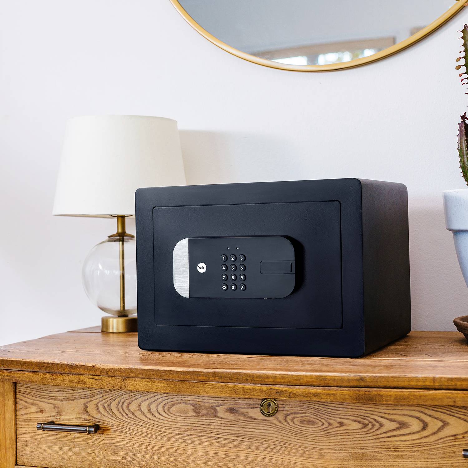 A black safe with an electronic combination lock sits on a wooden chest of drawers next to a lamp.