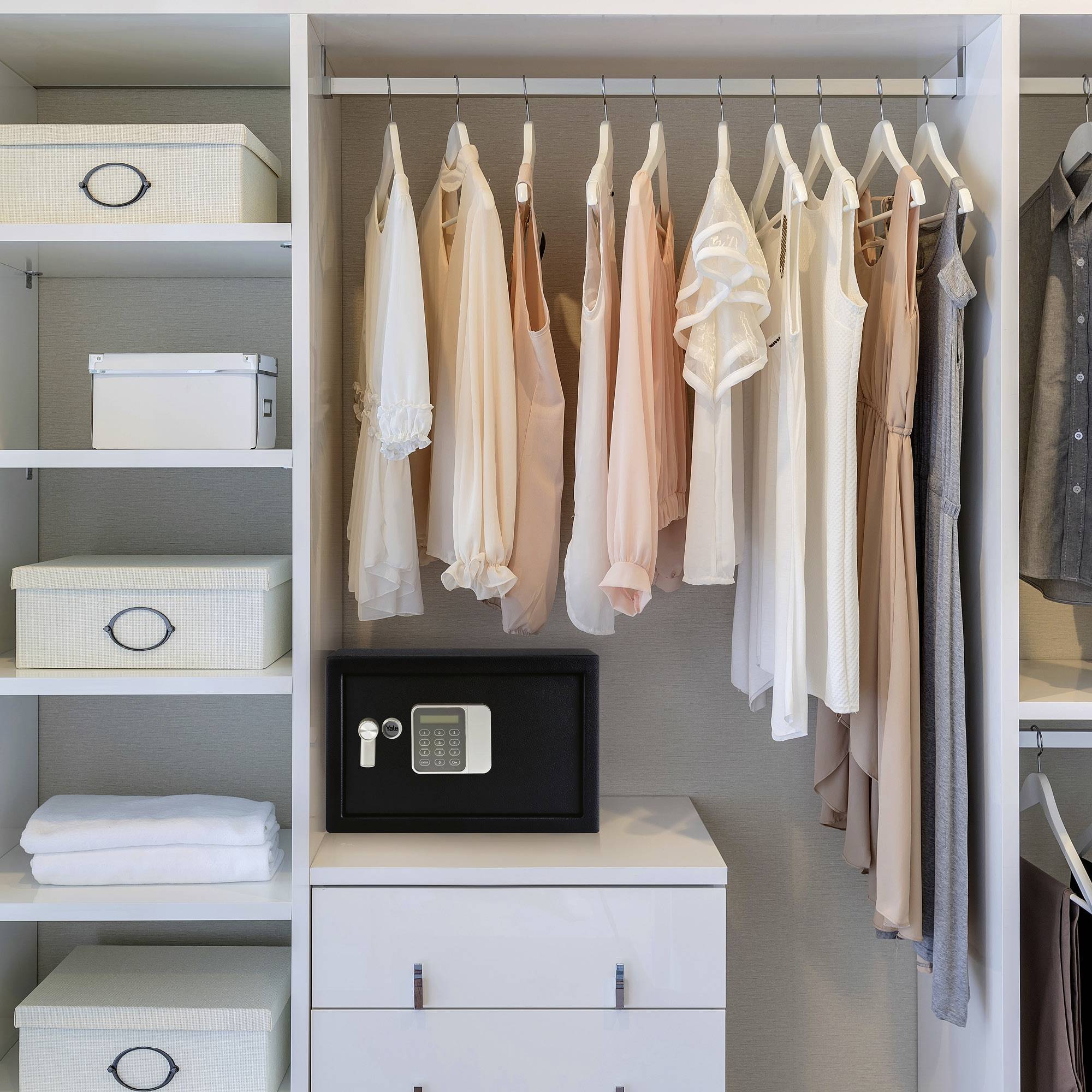 A tidy wardrobe with clothes in soft tones, a safe on the chest of drawers beneath, and baskets on the shelves beside it.