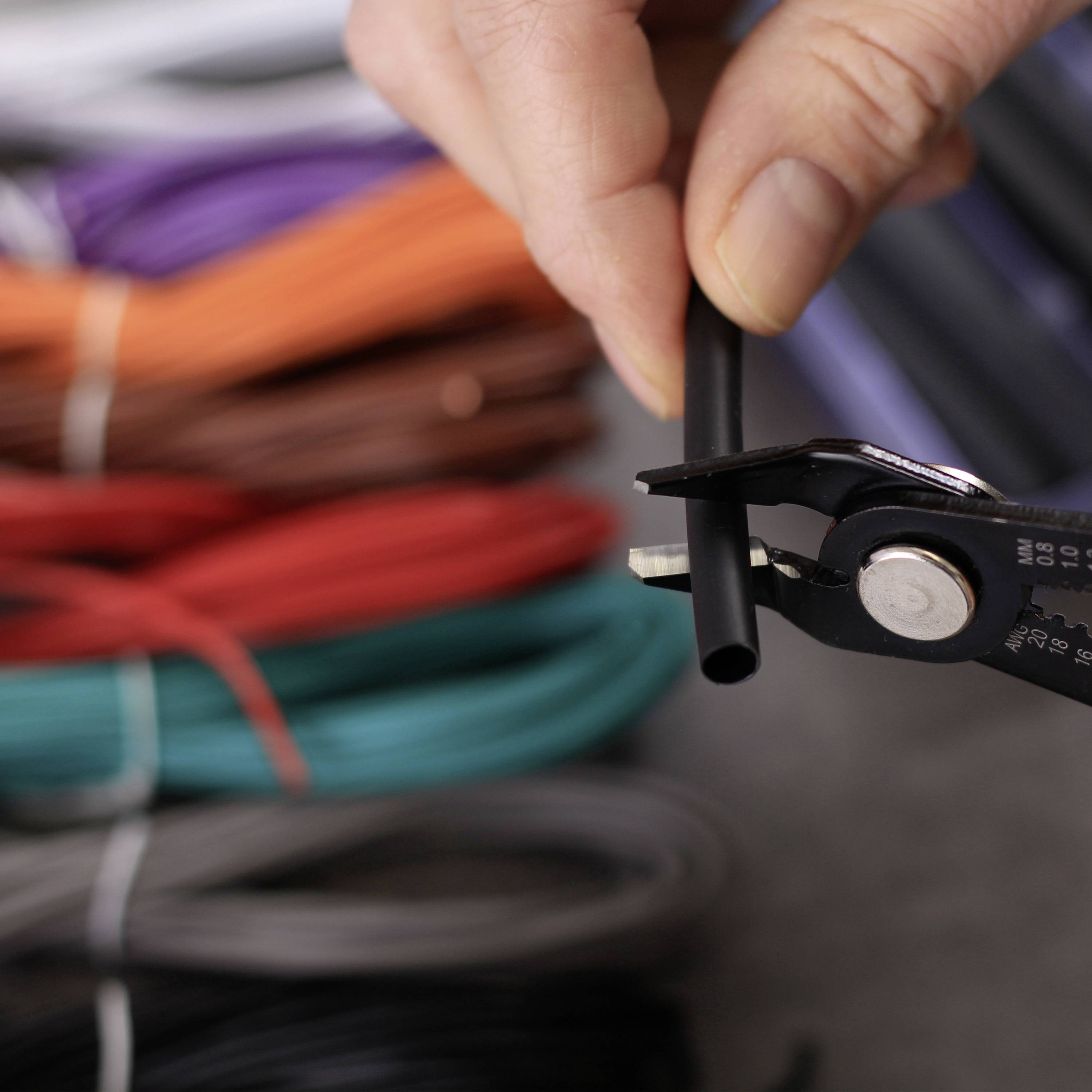 A hand is holding a side cutter and cutting a black cable. Colourful cables are visible in the background.