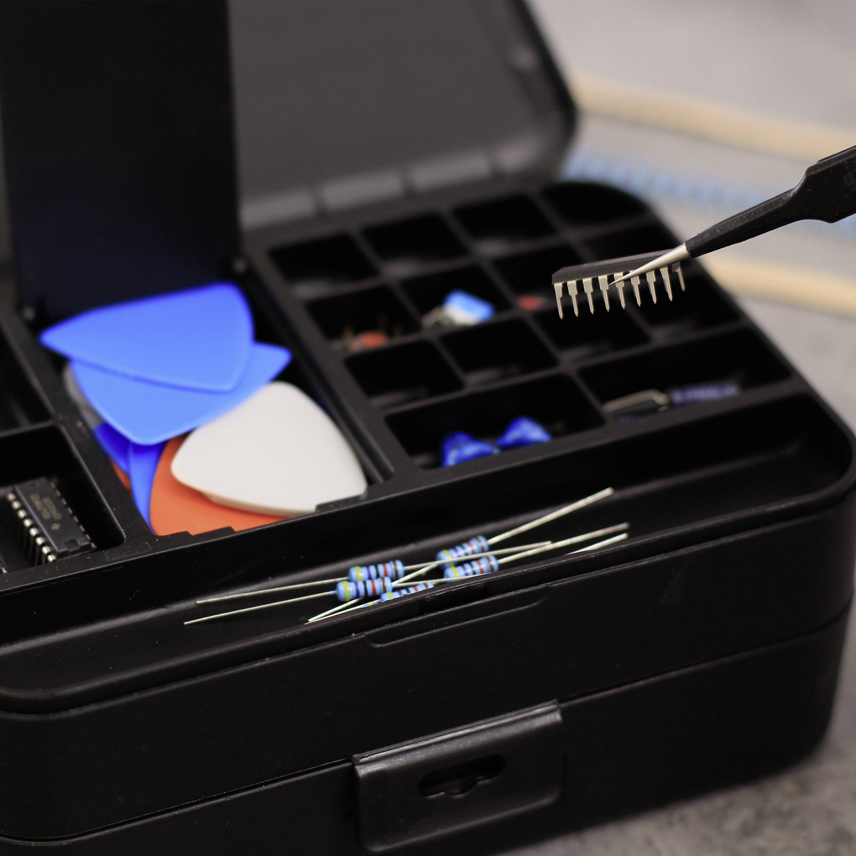 Tool case with guitar picks, resistors and other electronic components. A pair of tweezers is holding a small electronic part.
