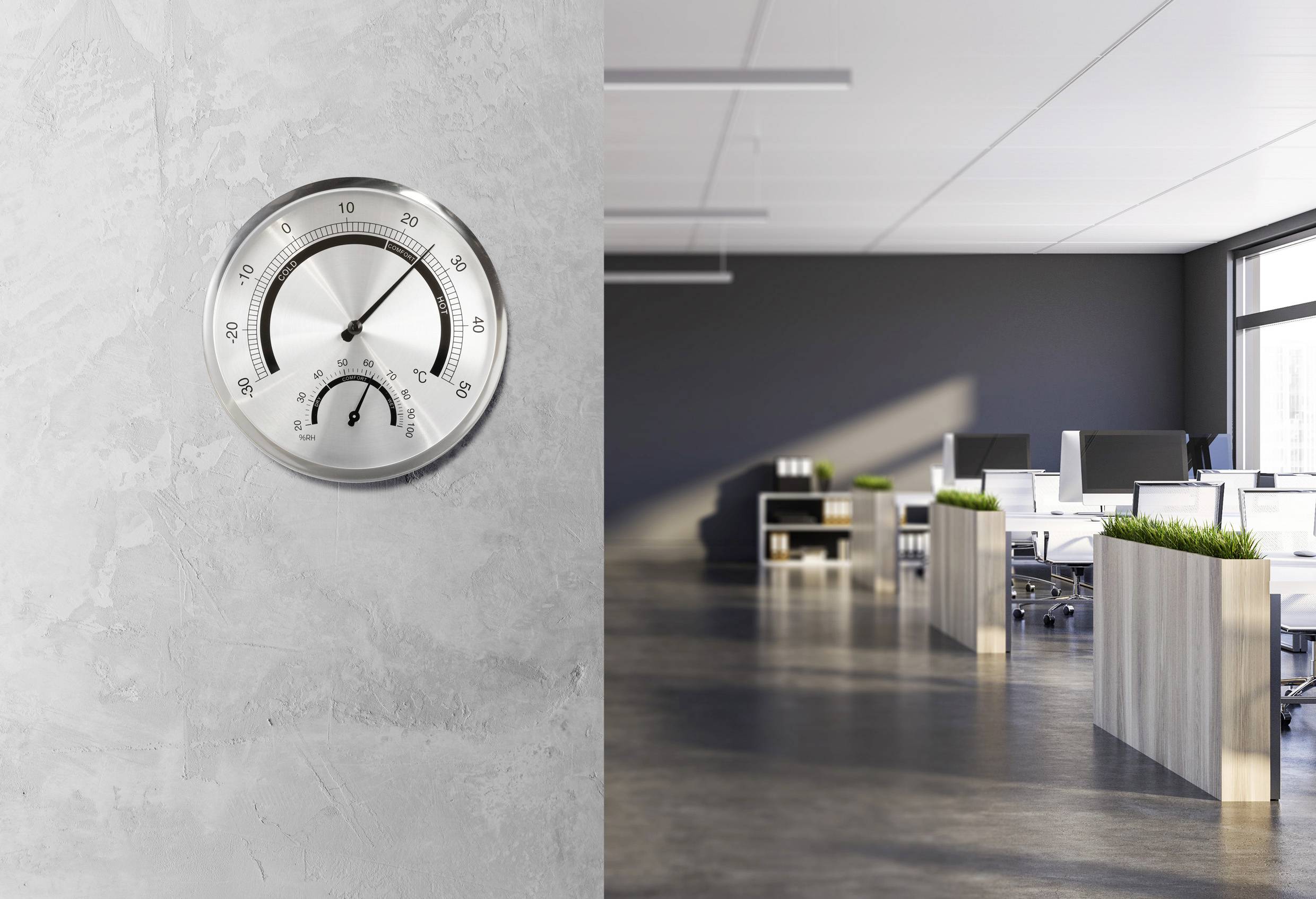 Bright office with modern desks and plants. A silver thermometer on the wall displays room temperature and humidity.