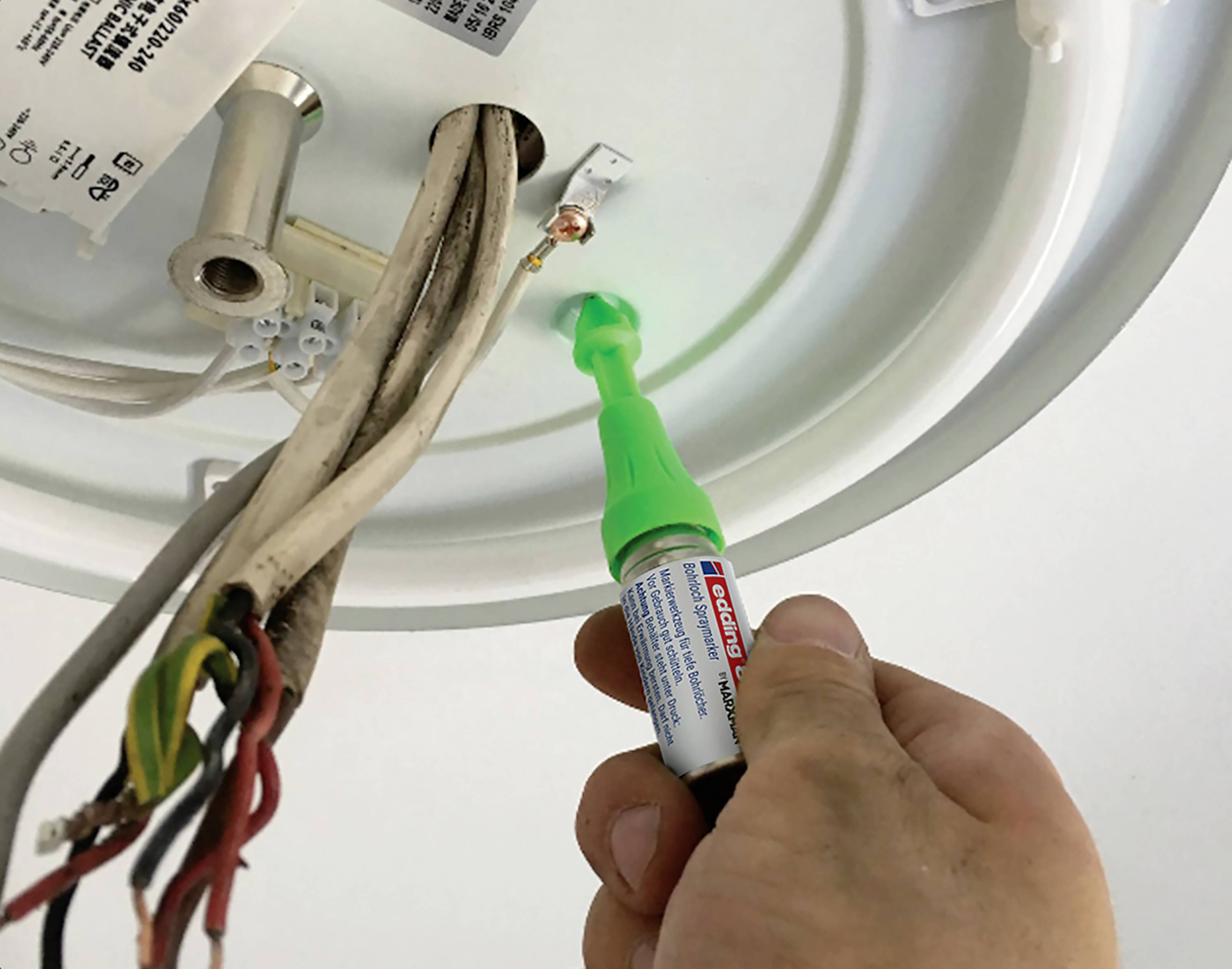 A hand holds a voltage tester against an electrical connection in a ceiling light. Several cables are visible. The device is checking the electrical current.