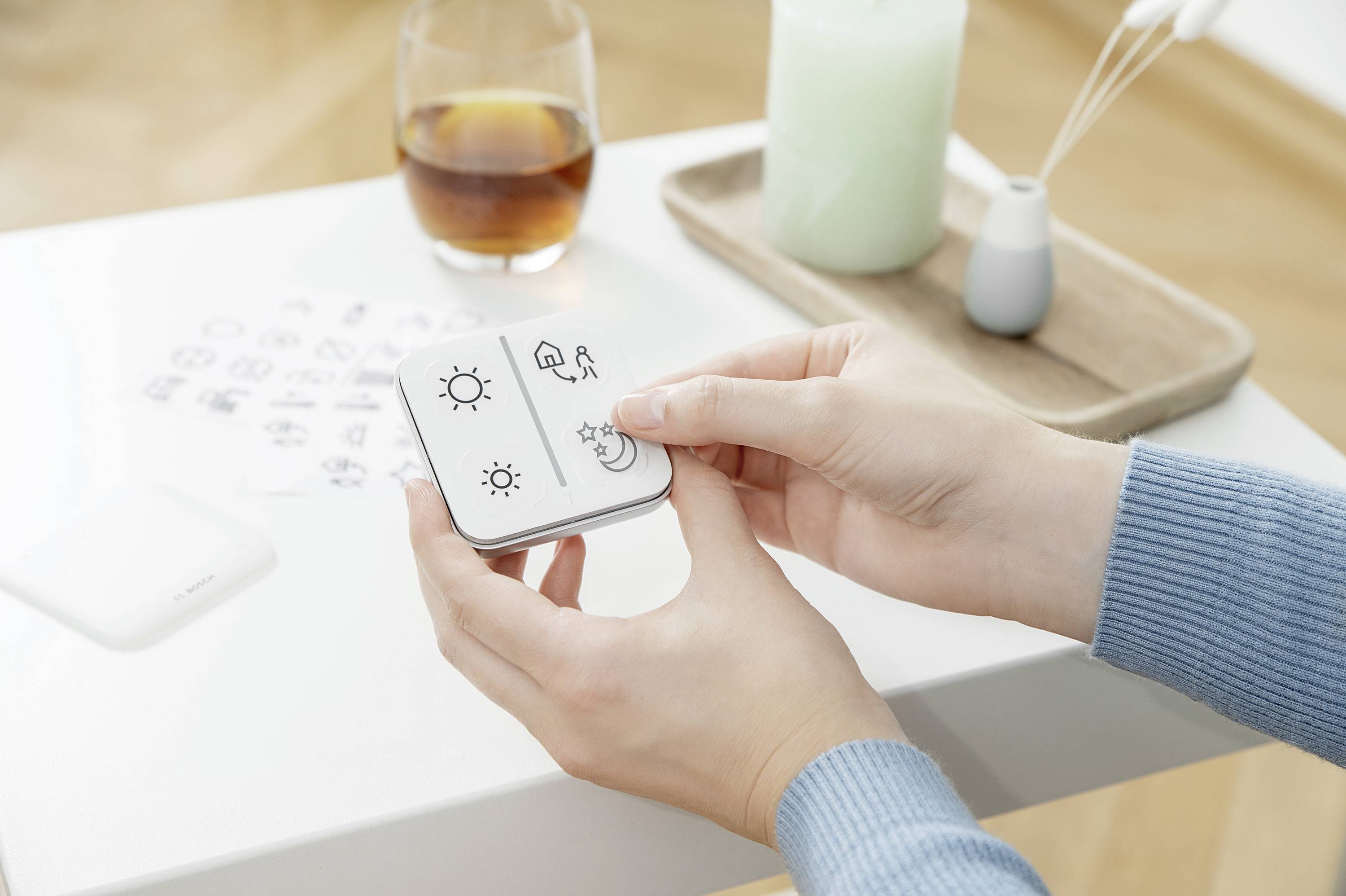 A person is holding a small device with symbols, including a sun and a moon. Next to them, a drink and a candle are placed on a table.