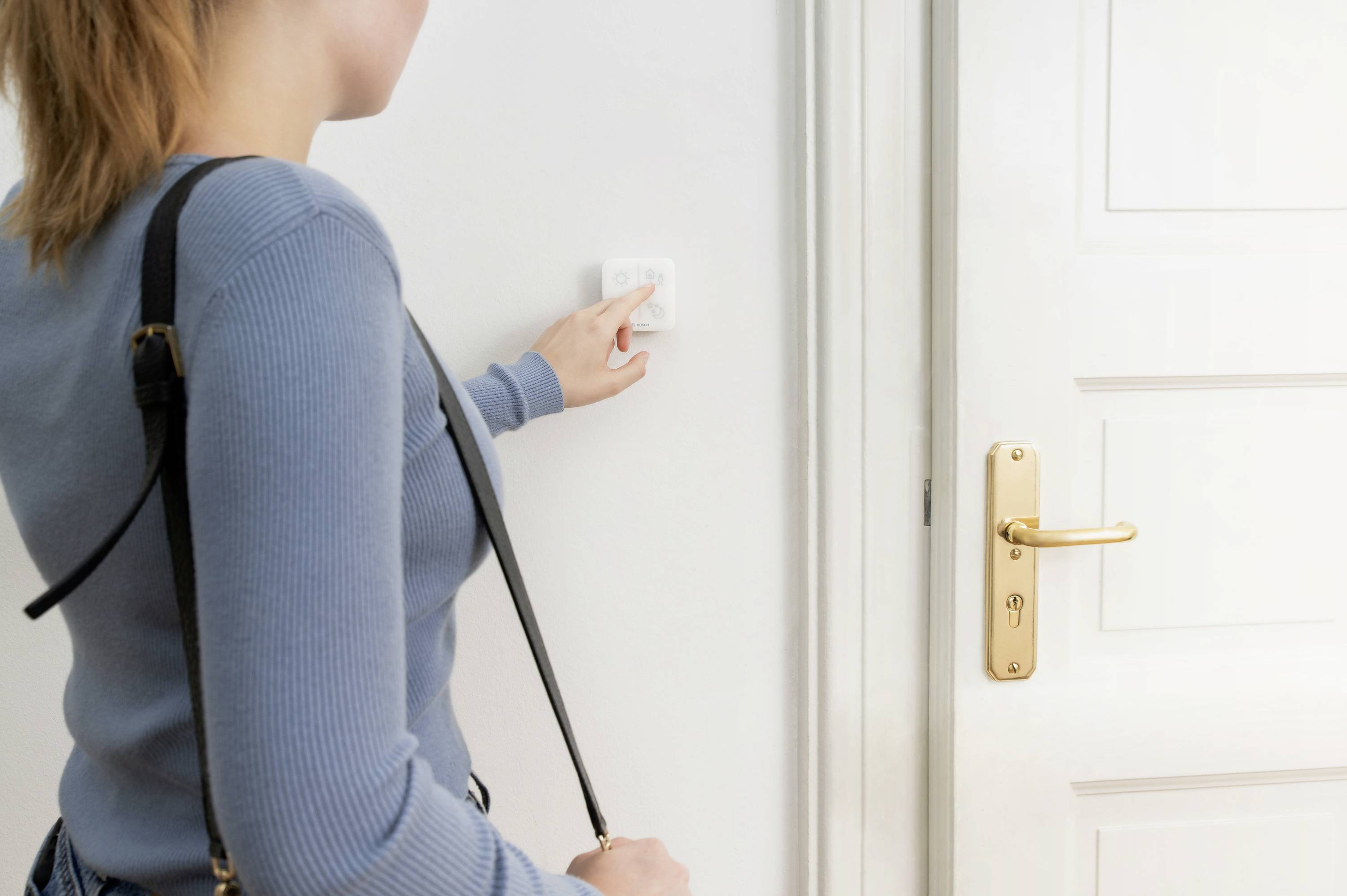 A person is pressing a light switch next to a white door. They are wearing a blue cardigan and a black shoulder bag.