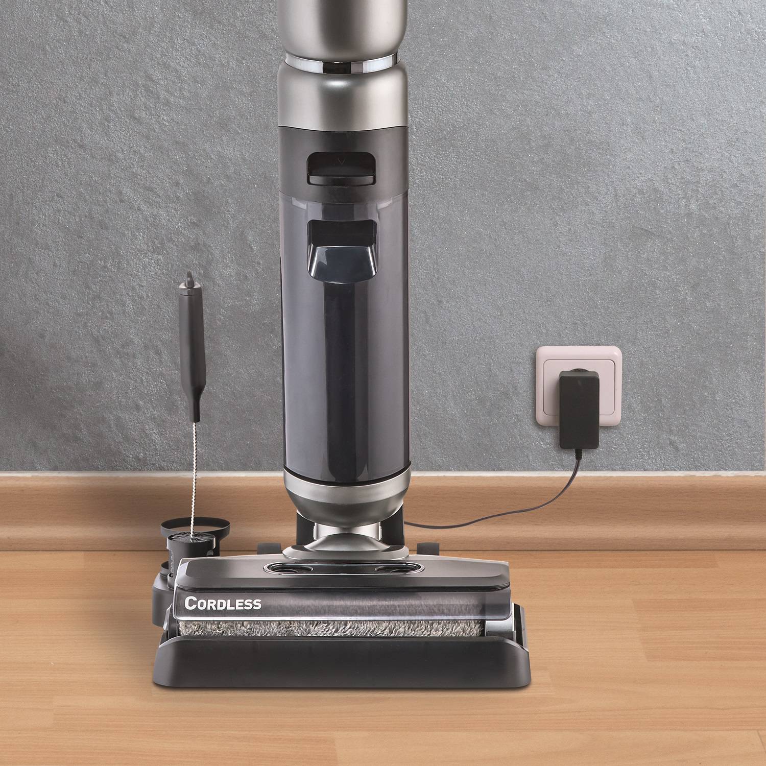 A cordless vacuum cleaner stands on a laminate floor, plugged into a socket. To the right of the hoover, the plug can be seen in the wall.