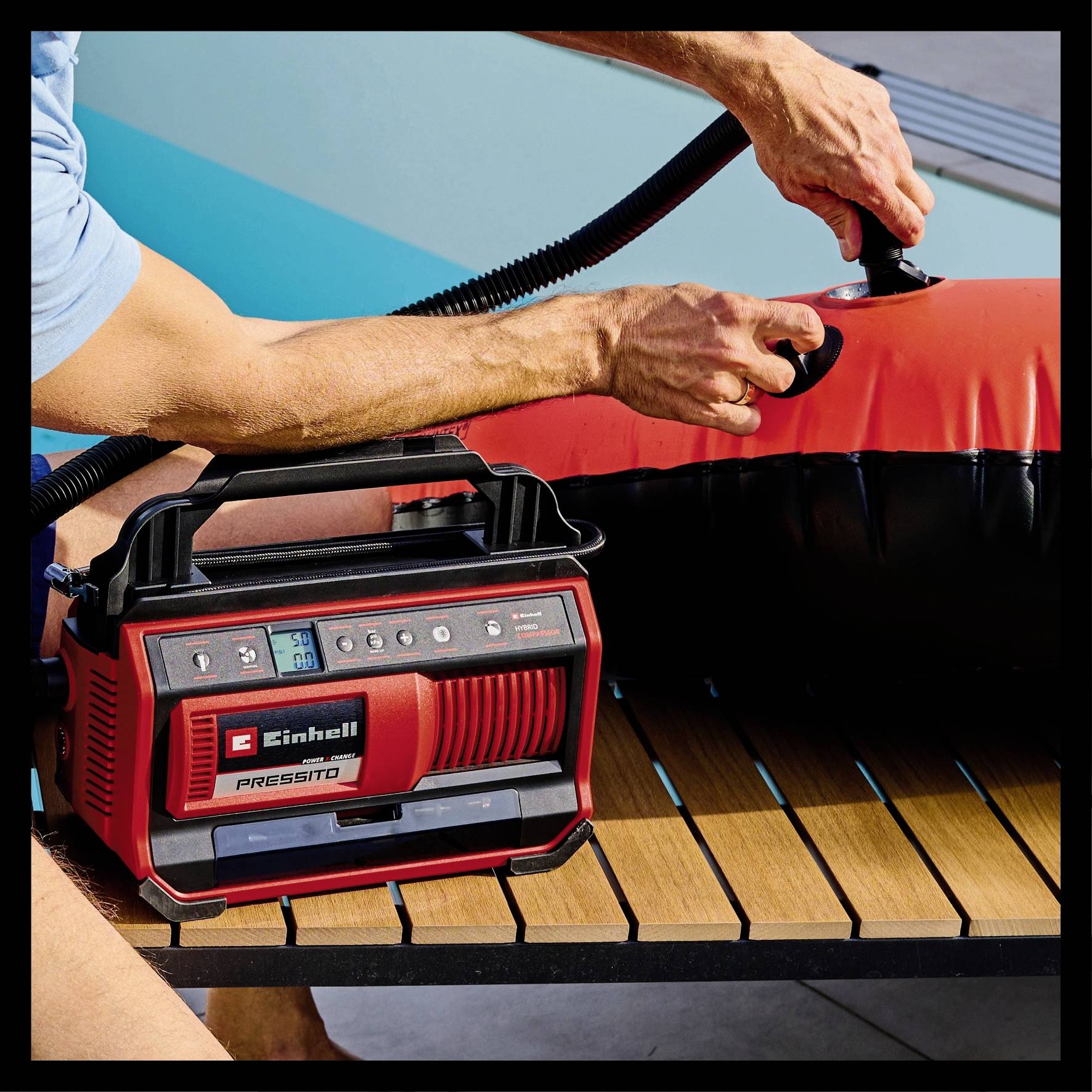 A man is inflating an air cushion at the poolside using a red, portable Einhell air compressor.