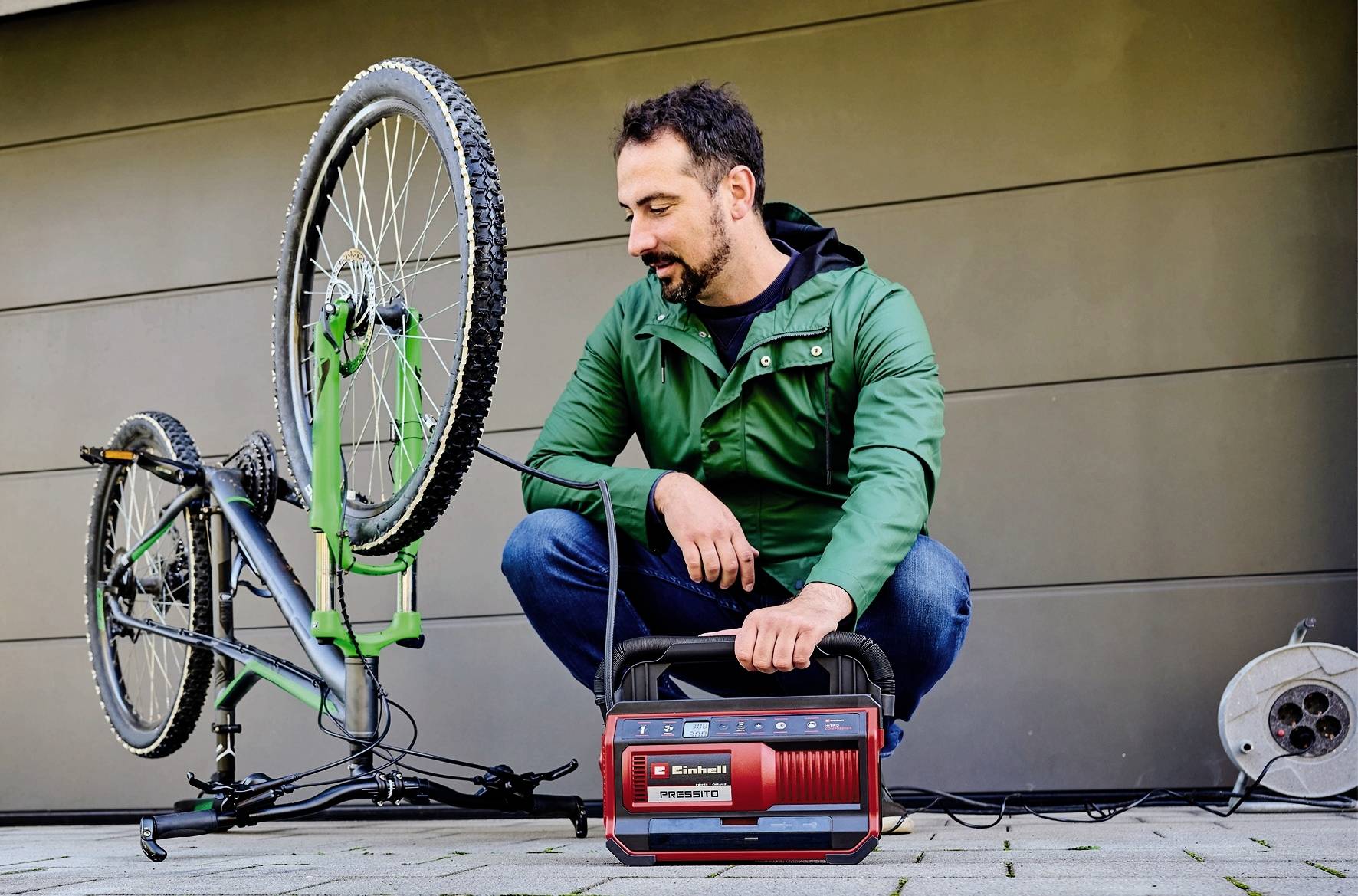 A man in a green anorak is repairing an upturned bicycle using a red electric tool.