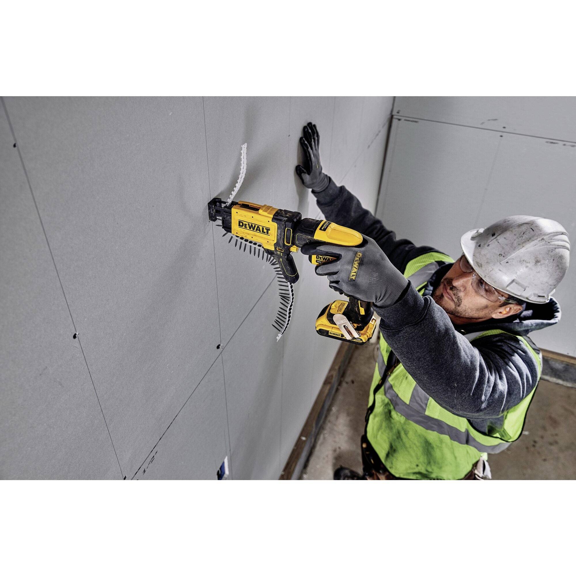 A worker in a hard hat and safety vest is screwing a plasterboard wall. He is using a yellow electric tool and wearing gloves.