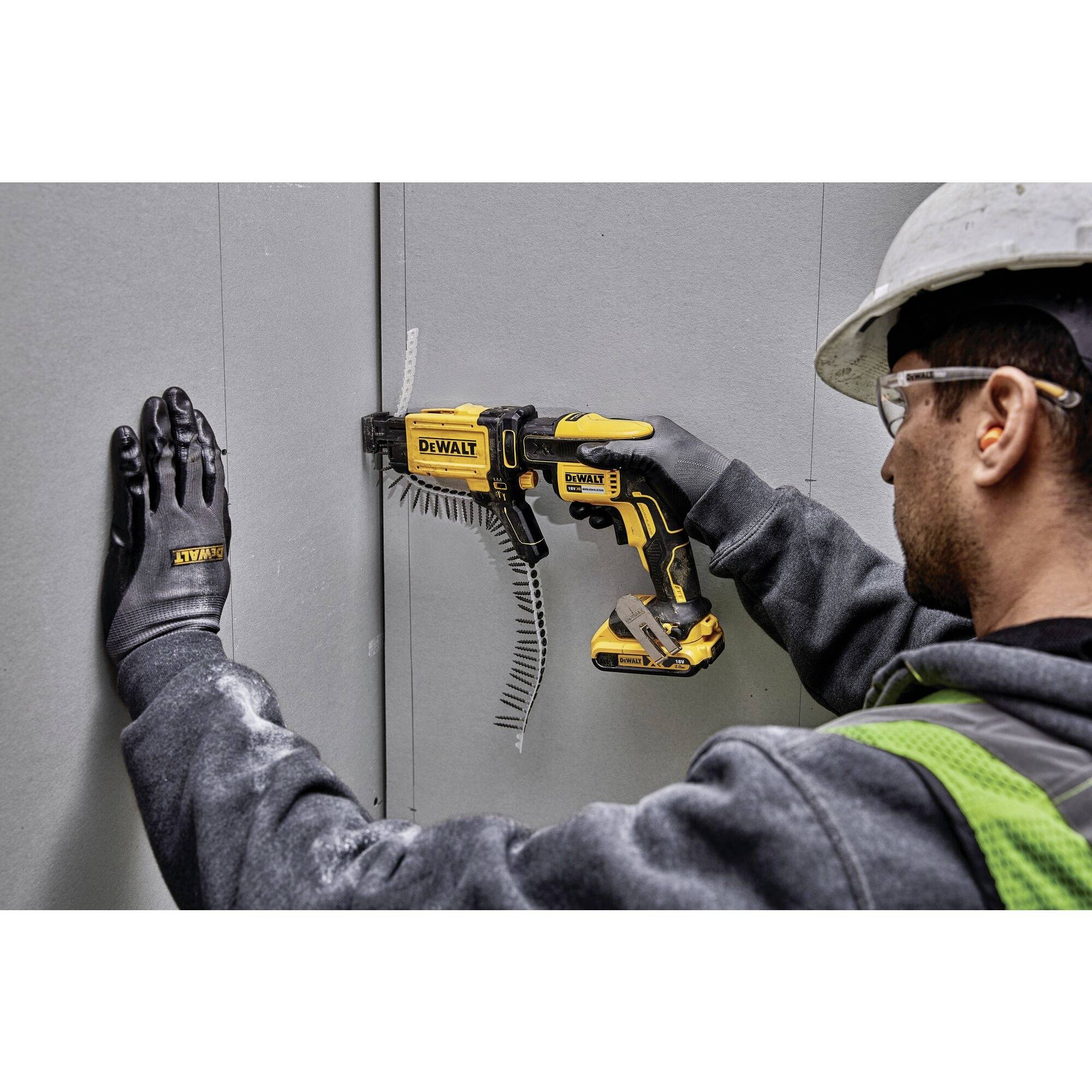 A person wearing protective equipment is using a yellow power tool to secure a screw into a grey wall.