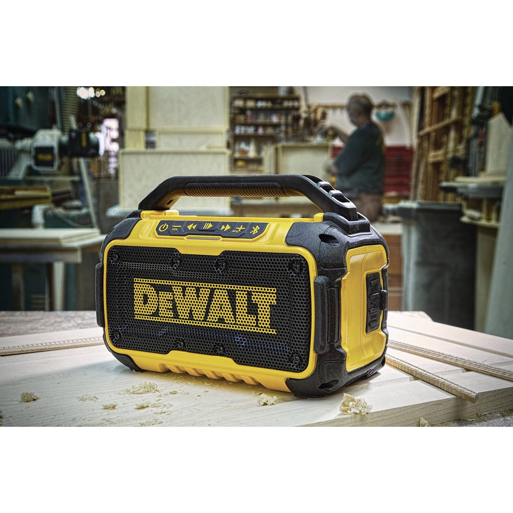 Portable, yellow speaker with 'DEWALT' logo sitting on a wooden workbench in a workshop. Tools are visible in the background.
