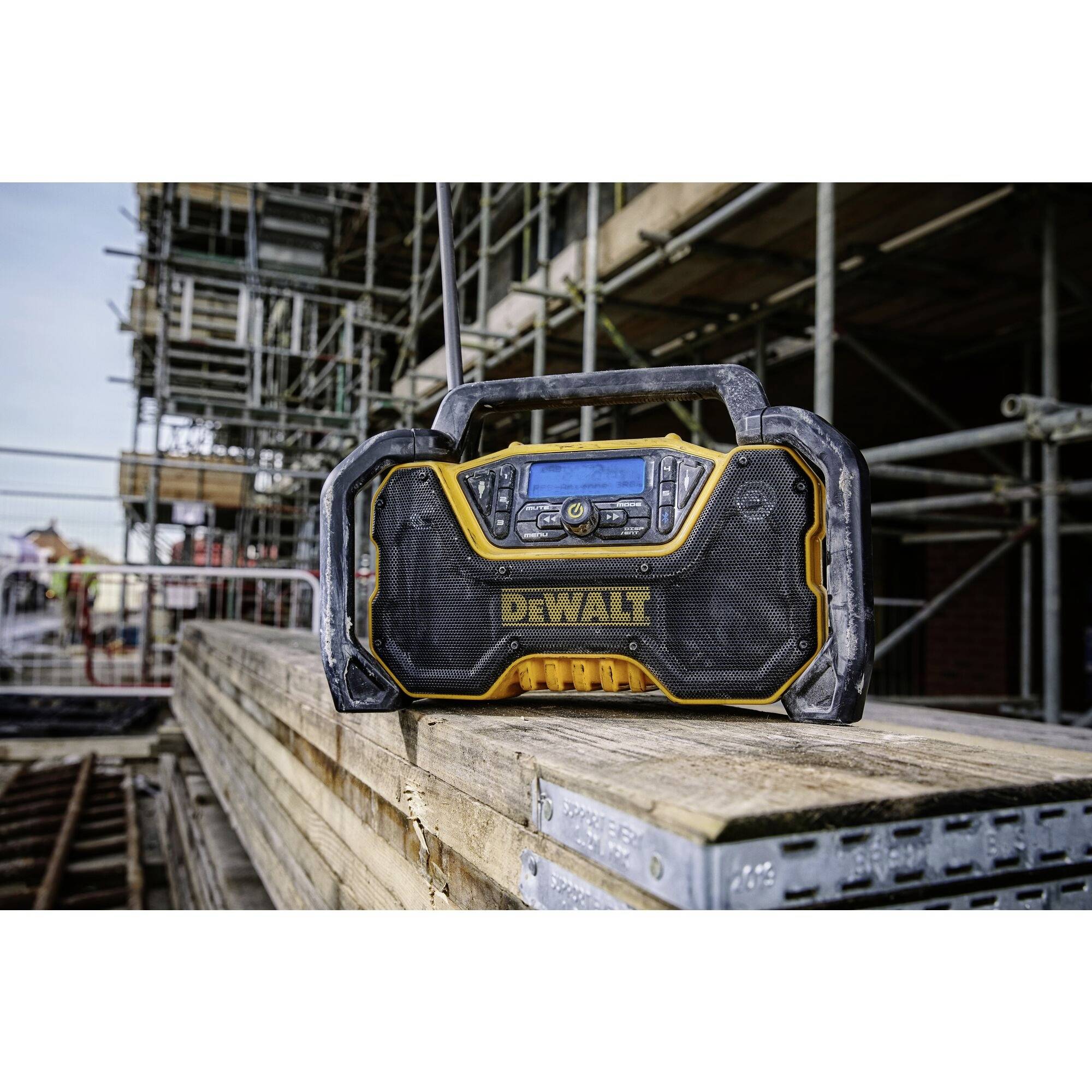 A robust site radio on a pile of timber, with scaffolding and a construction site in the background.