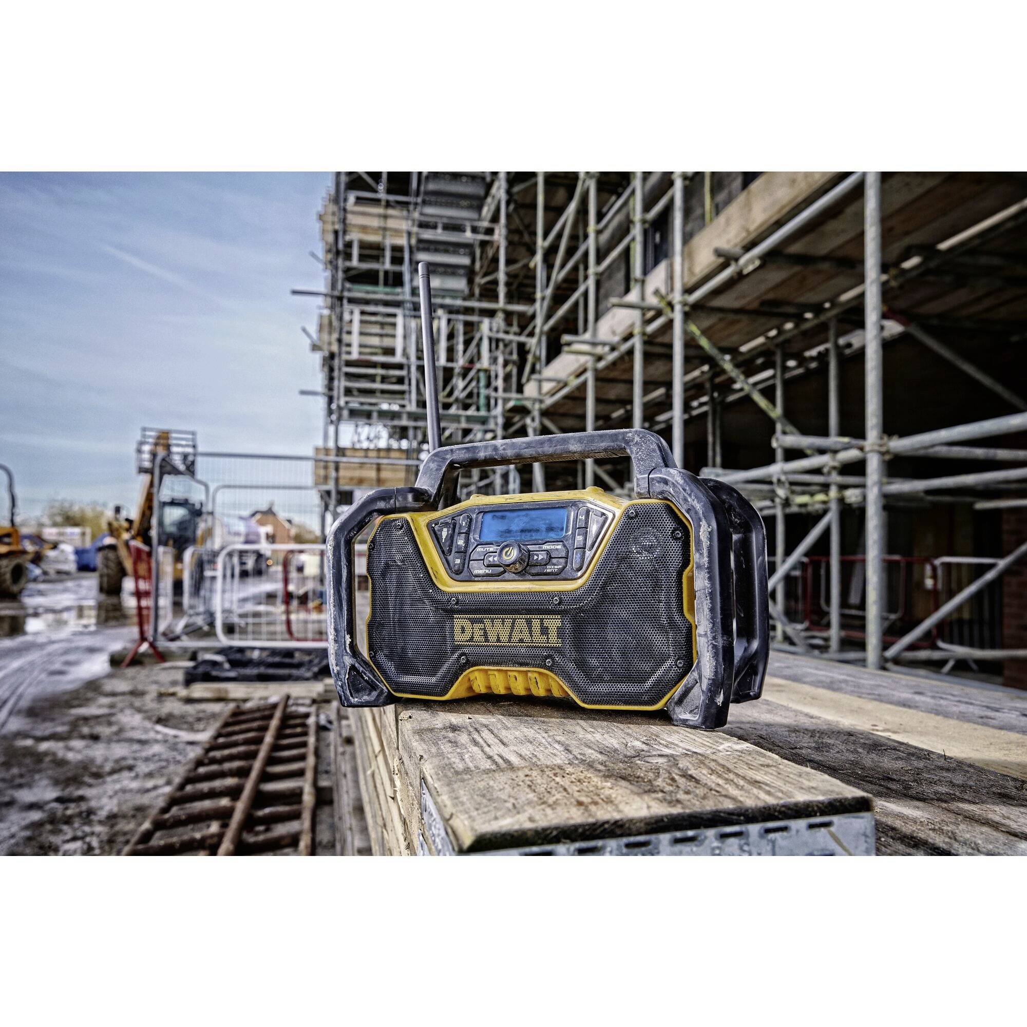 Construction site radio on a wooden table with a building site in the background, scaffolding and building materials, demonstrating robust design under construction site conditions.