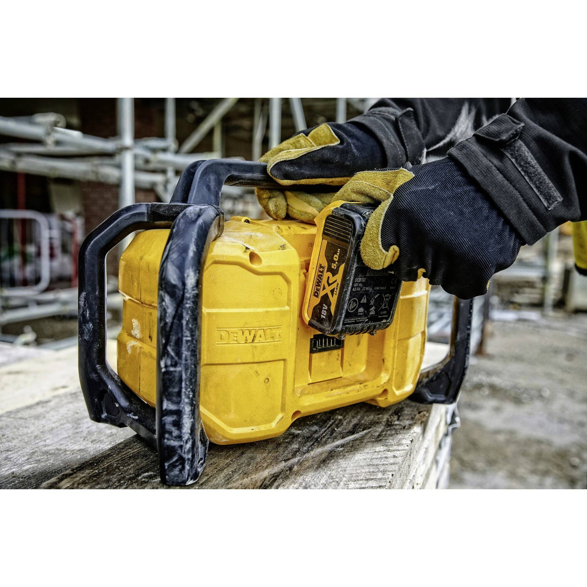 Two gloved hands are operating a yellow construction site radio. Scaffolding and parts of a construction site are visible in the background.