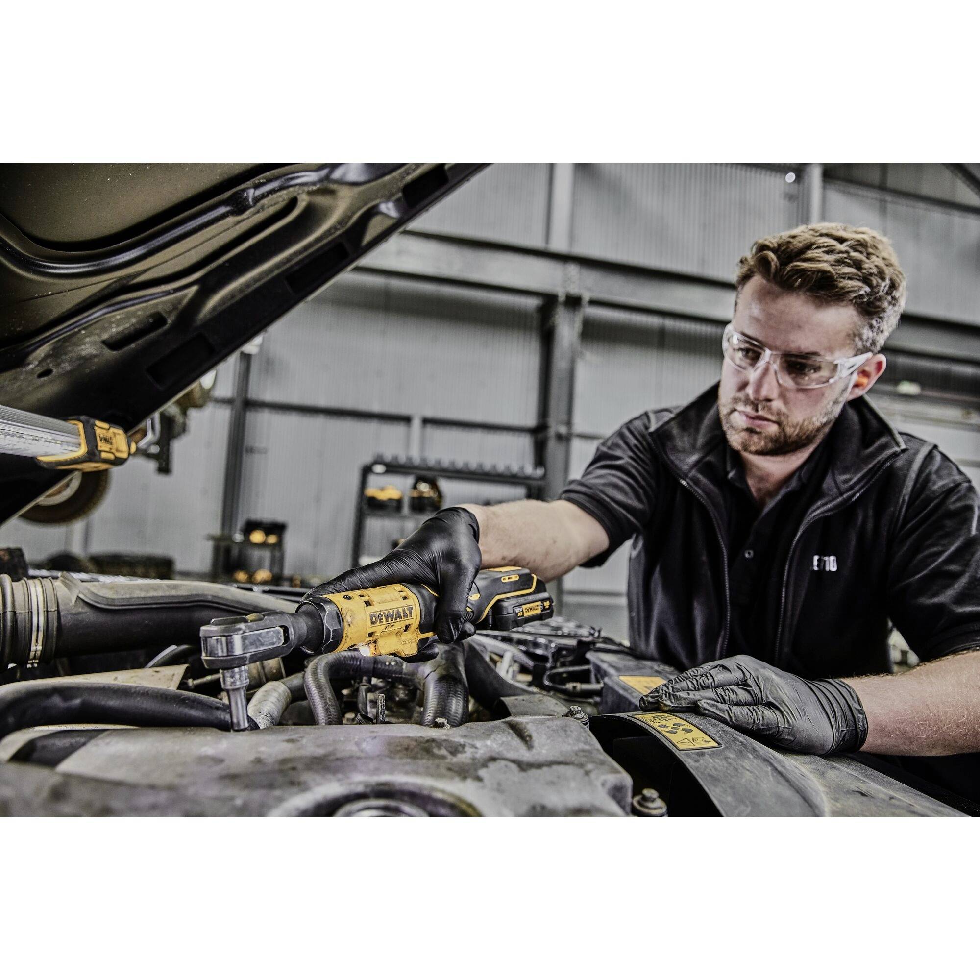 A mechanic wearing safety goggles and gloves is working with an electric tool in an open engine compartment within a workshop environment.